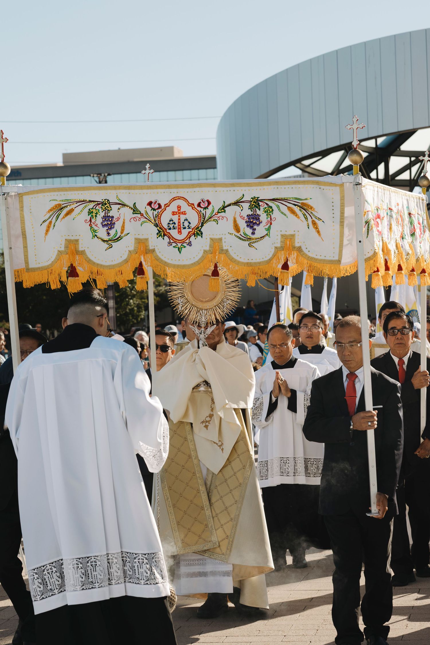 Religious procession with ornate canopy and clergy in white robes walking among gathered crowd.