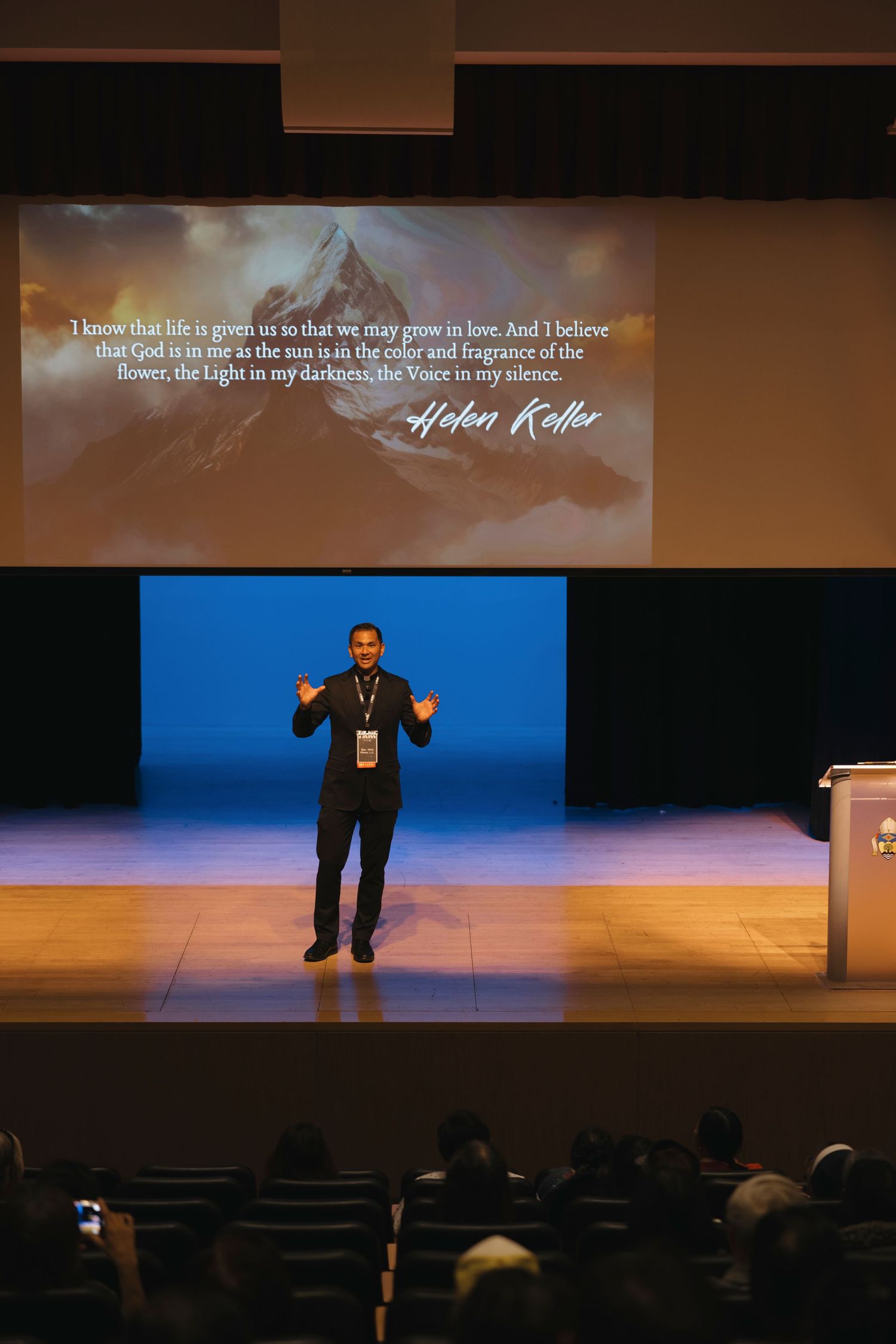A speaker presents on stage with projected text visible on a large screen in an auditorium setting.