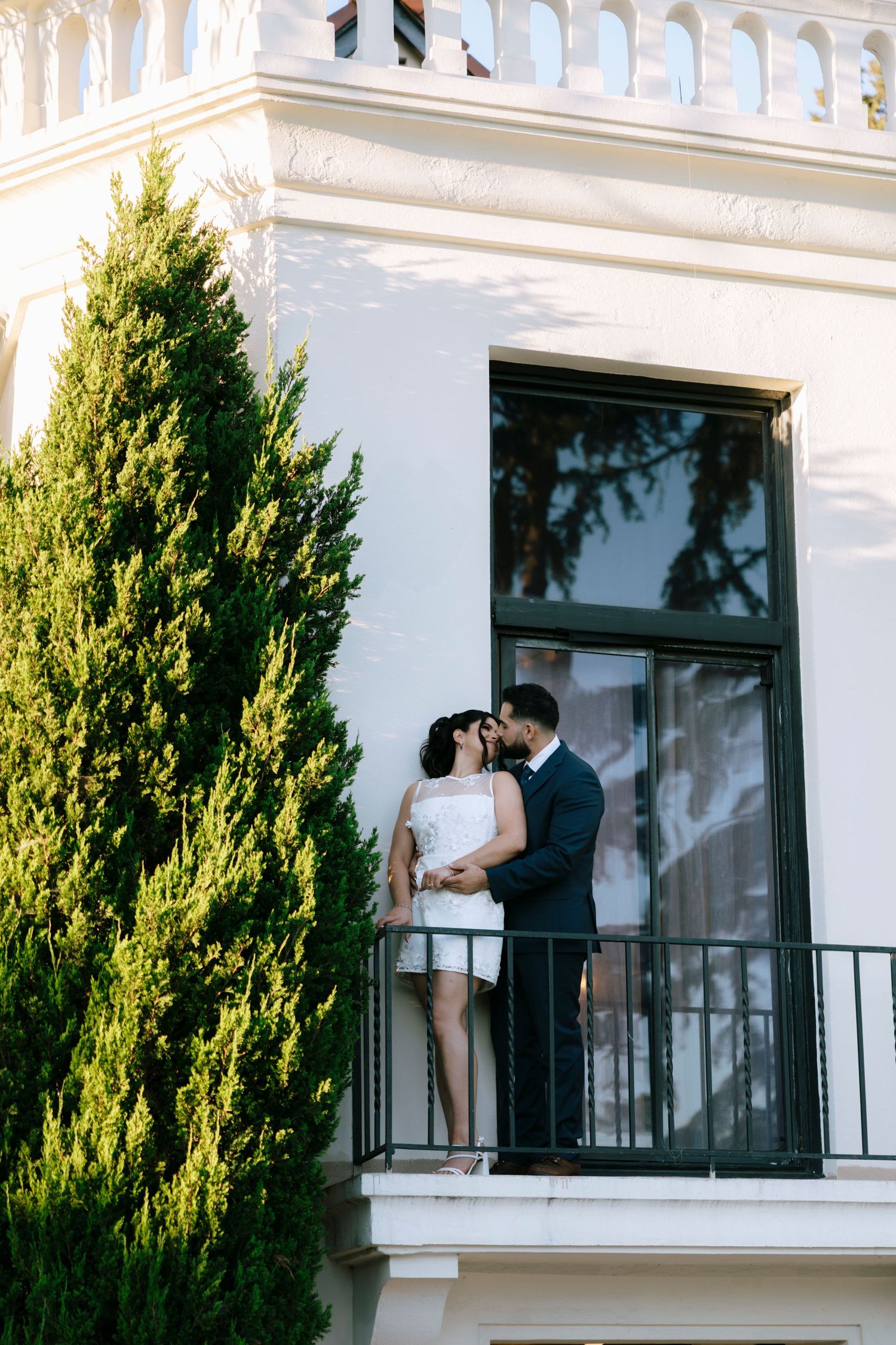 A couple embraces on an elegant white balcony with green cypress trees in view.