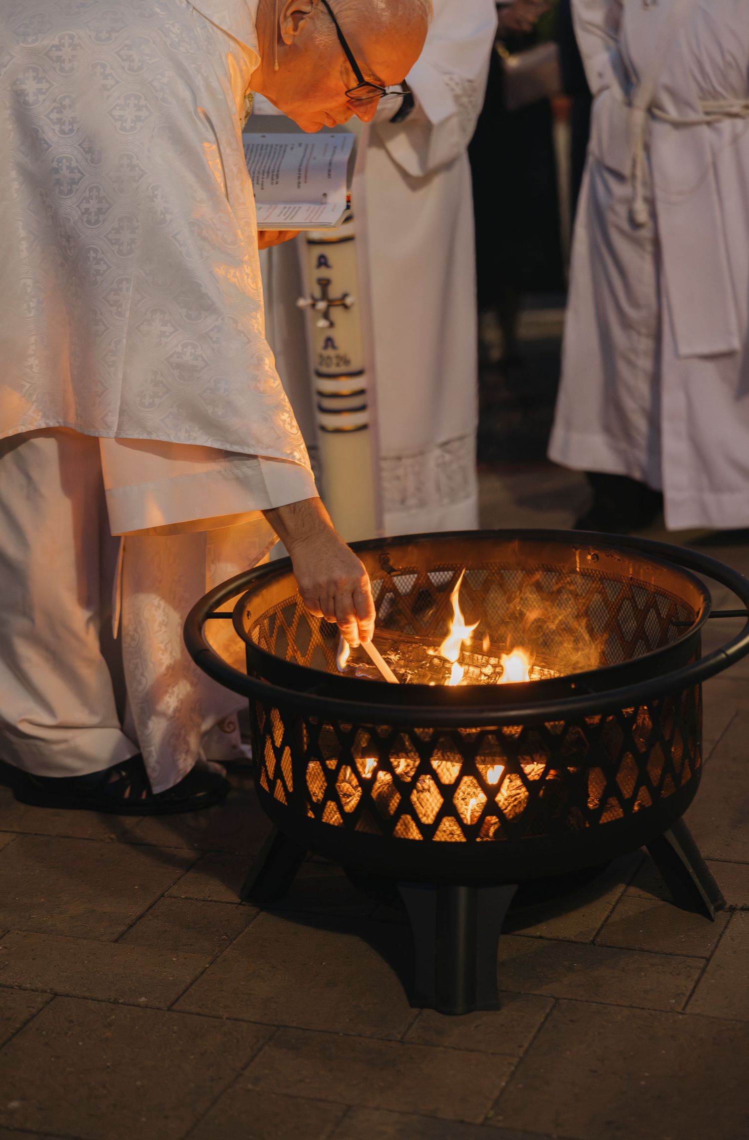 Priest in white robes lighting a fire in a black metal outdoor fire pit during a religious ceremony.