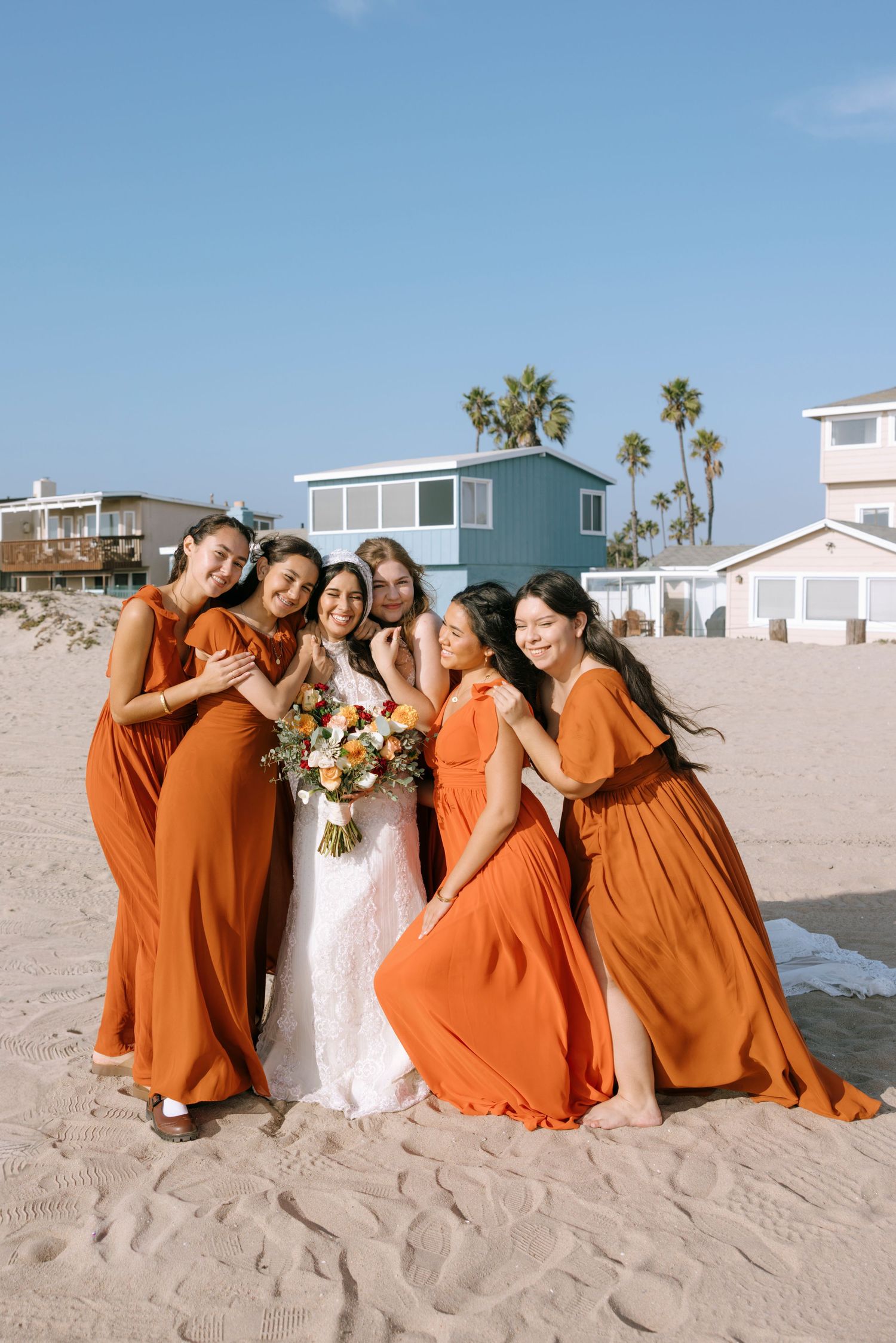 Bridesmaids in vibrant orange dresses pose playfully with the bride on a sunny beach.