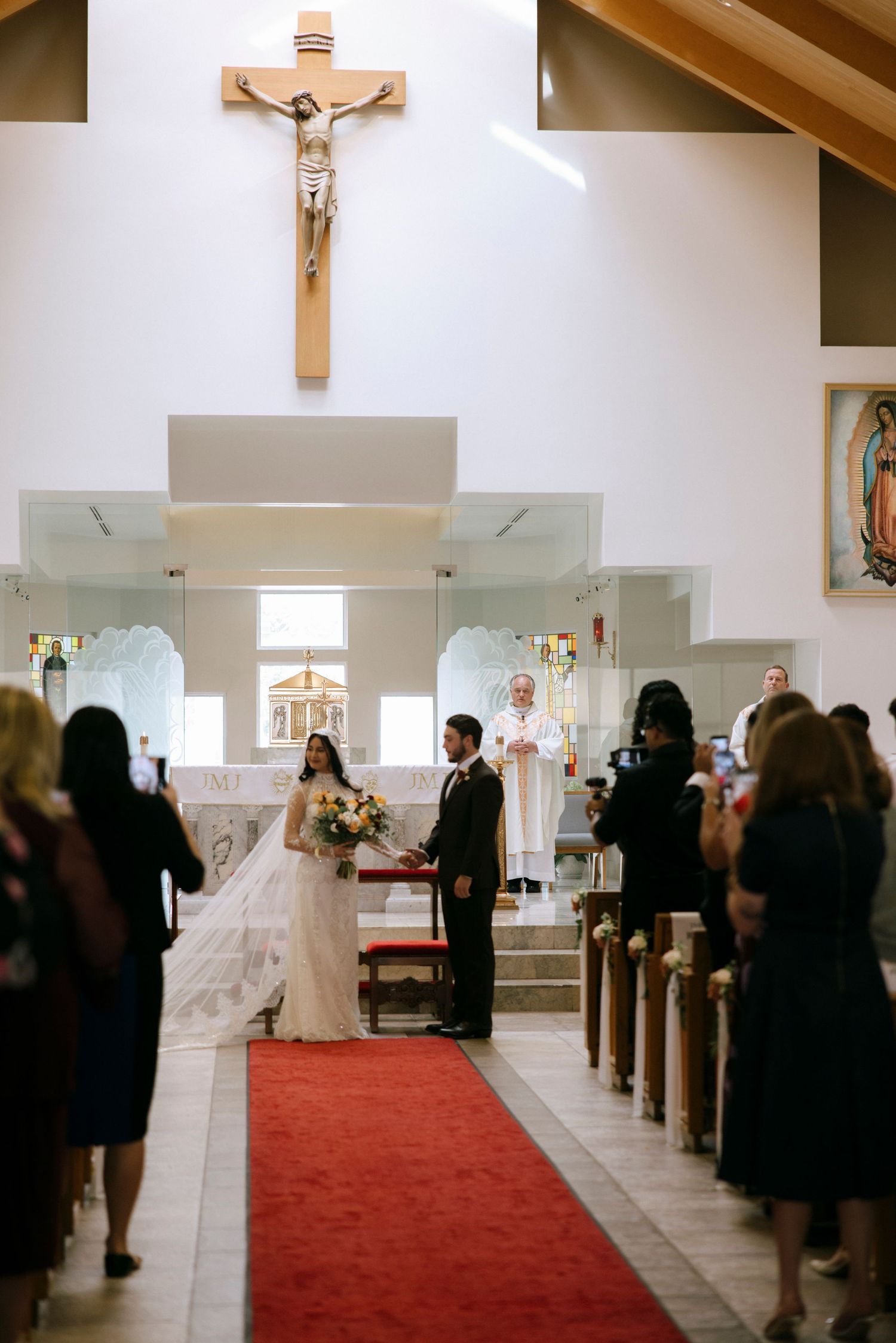 A Catholic church wedding ceremony with a red carpet aisle and wooden pews filled with guests.