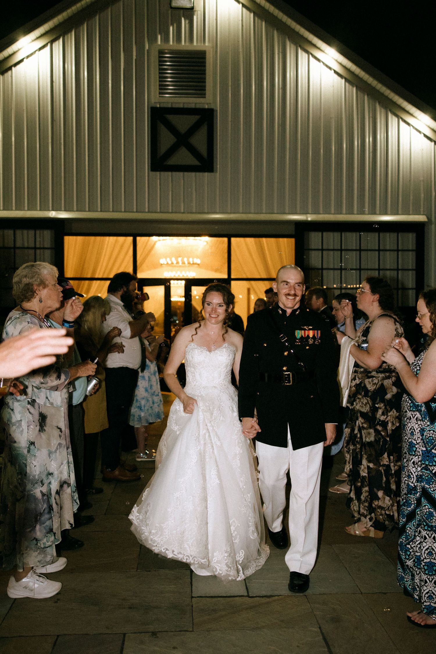 Bride in white gown and groom in military dress uniform exit wedding venue while guests celebrate.