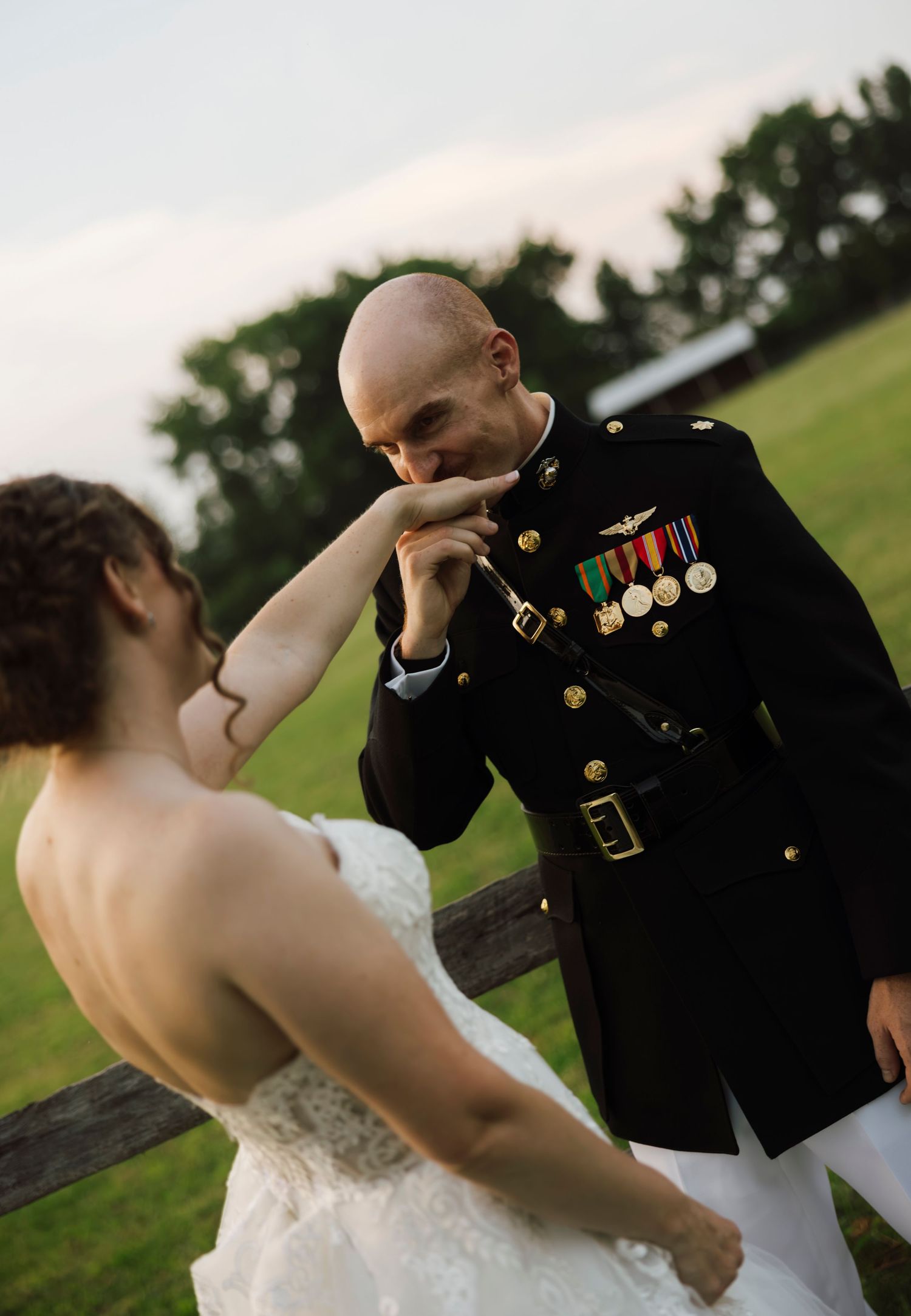 Marine in dress uniform kisses bride's hand during outdoor wedding ceremony near rustic fence.