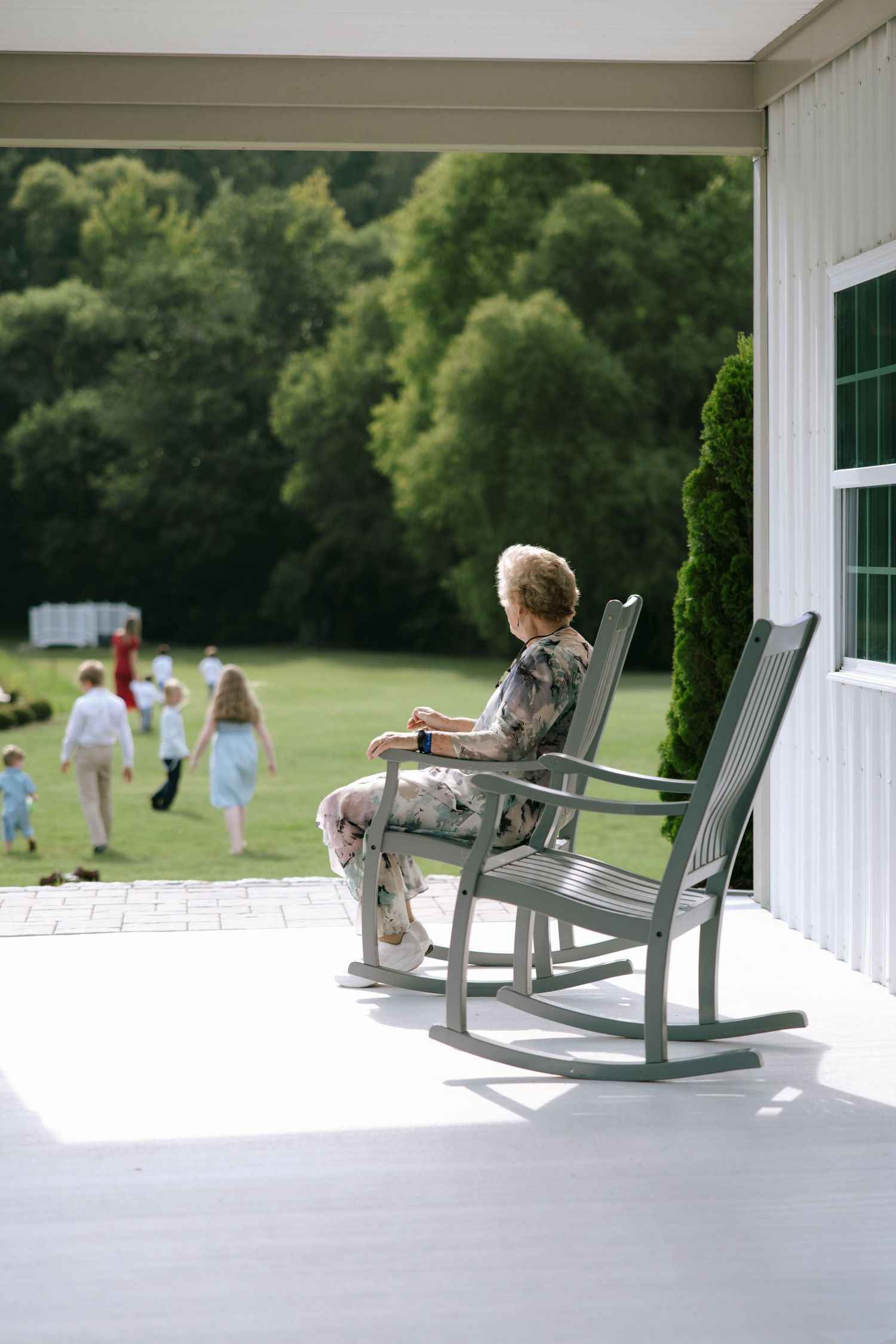 Elderly woman sitting in gray rocking chair on white porch, watching children play on green lawn with trees in background.