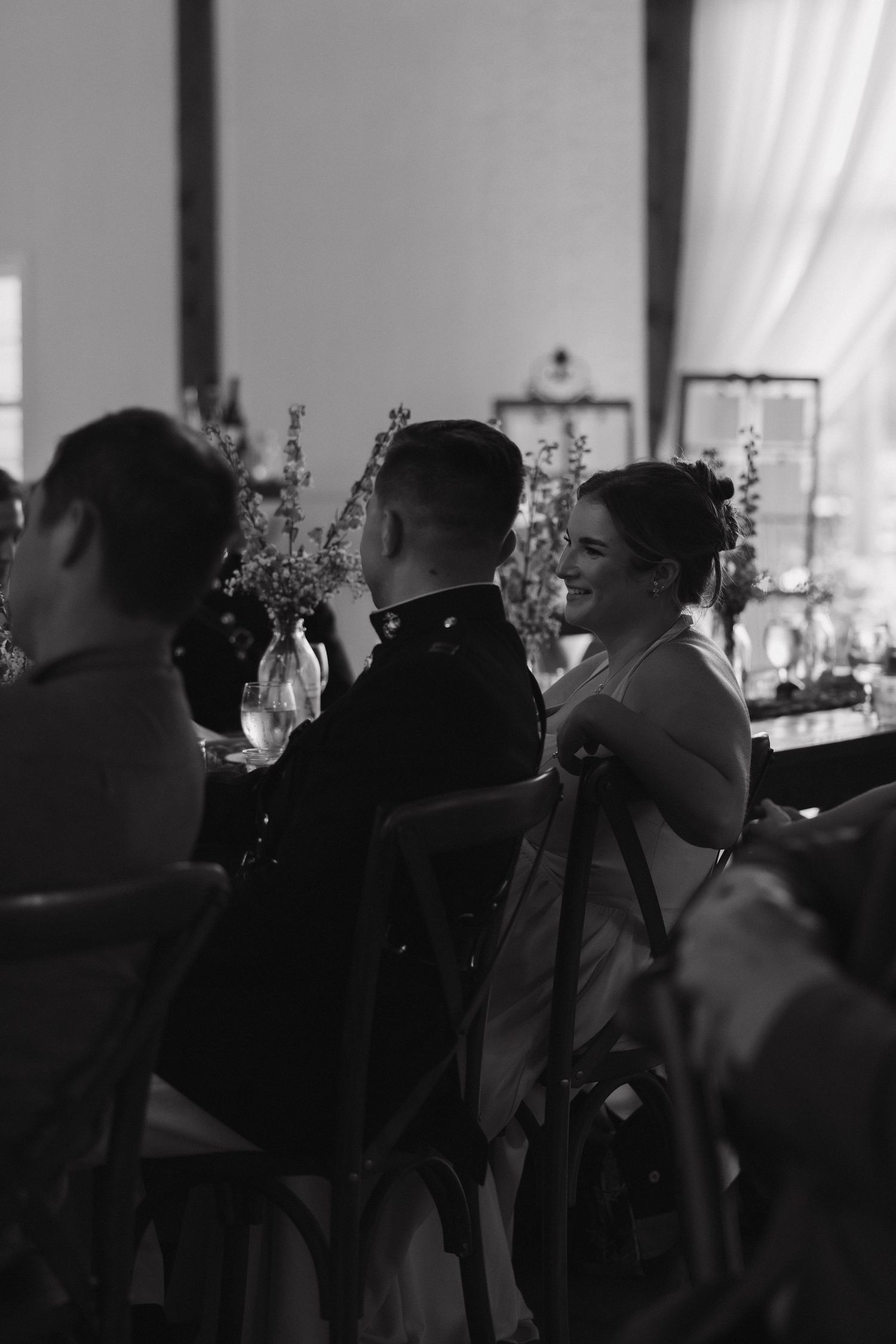 Black and white photo of a smiling bride in a white dress seated beside a groom in military uniform at their wedding reception.