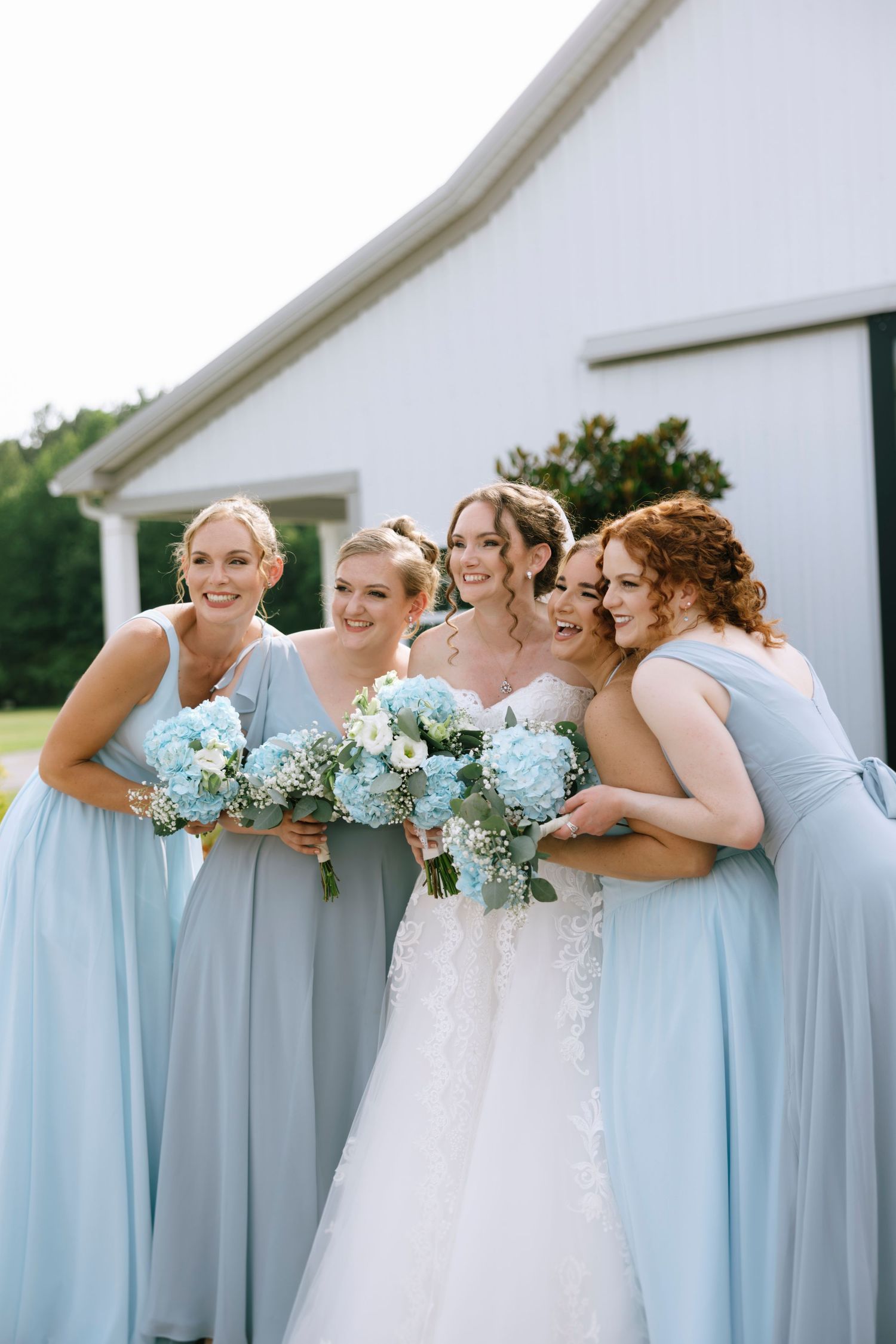 Bride in white lace gown poses with four bridesmaids in light blue dresses holding matching floral bouquets.