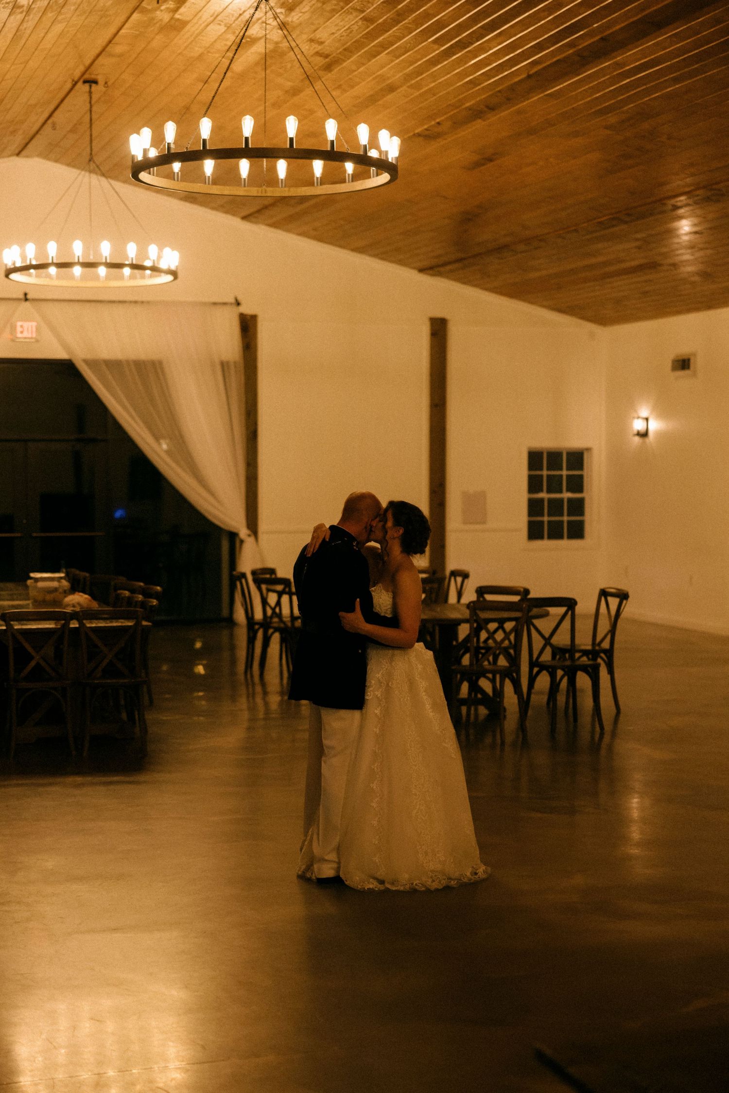 A bride and groom share their first dance under a rustic chandelier in an elegant barn wedding venue.