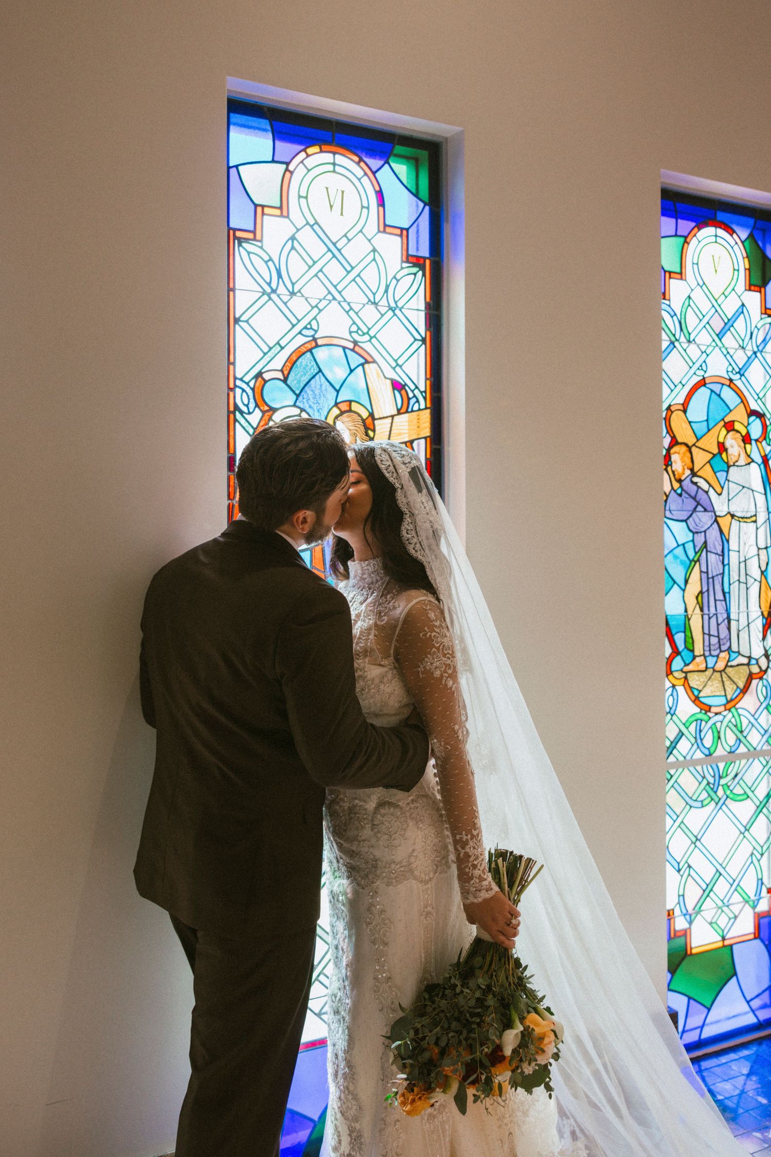 A couple shares their first kiss as newlyweds in front of a beautiful stained glass window in a church.