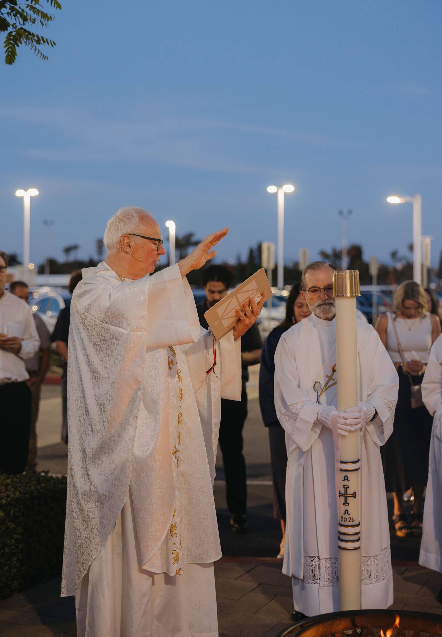 Priest in white vestments raises hand in blessing during outdoor Easter Vigil ceremony at dusk.