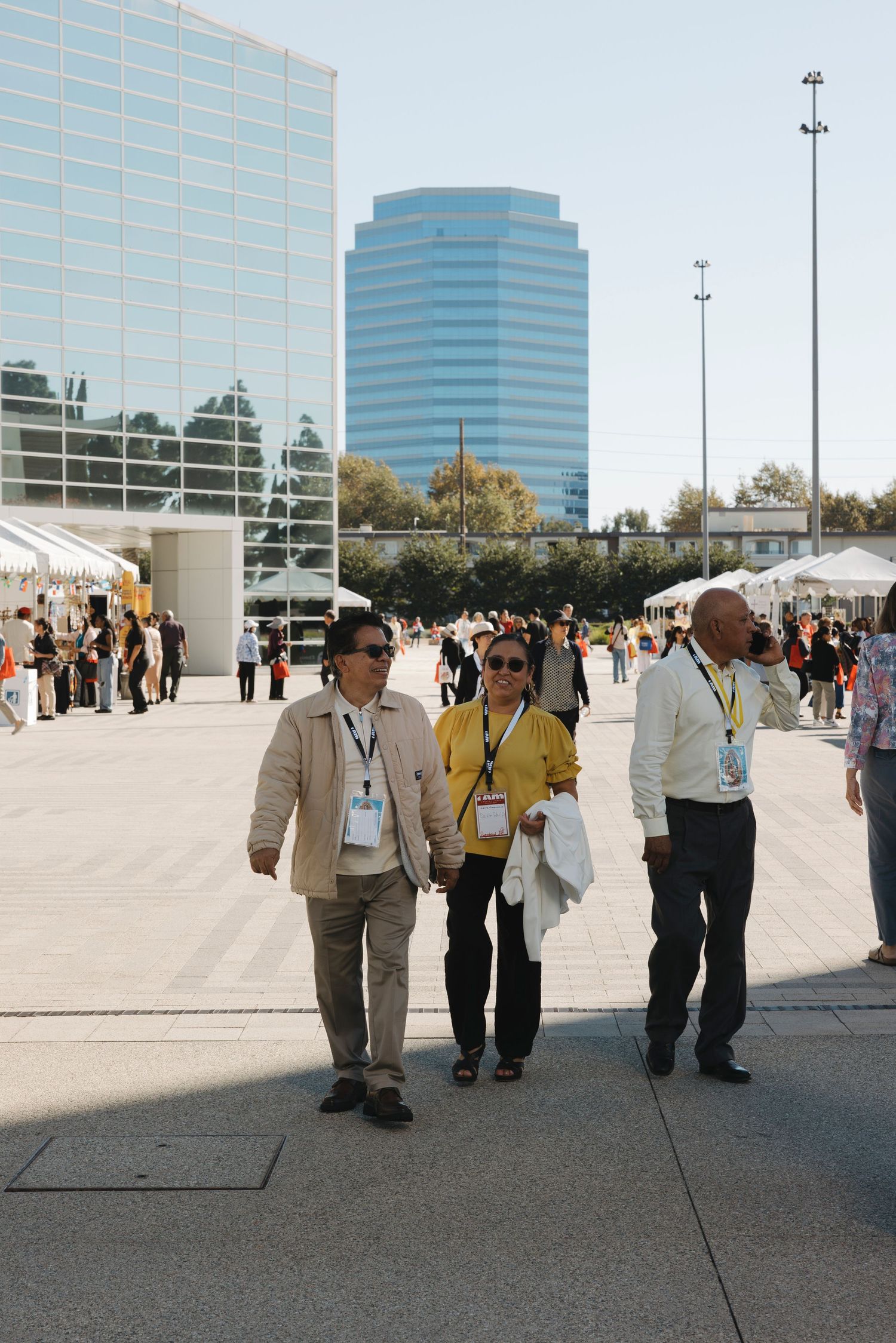 Attendees with lanyards walk through an outdoor event space with modern buildings in background.