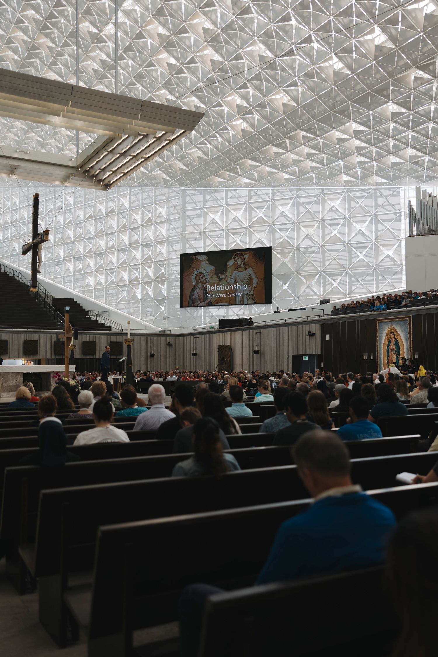 A modern church sanctuary with geometric ceiling patterns and large screens filled with attendees.