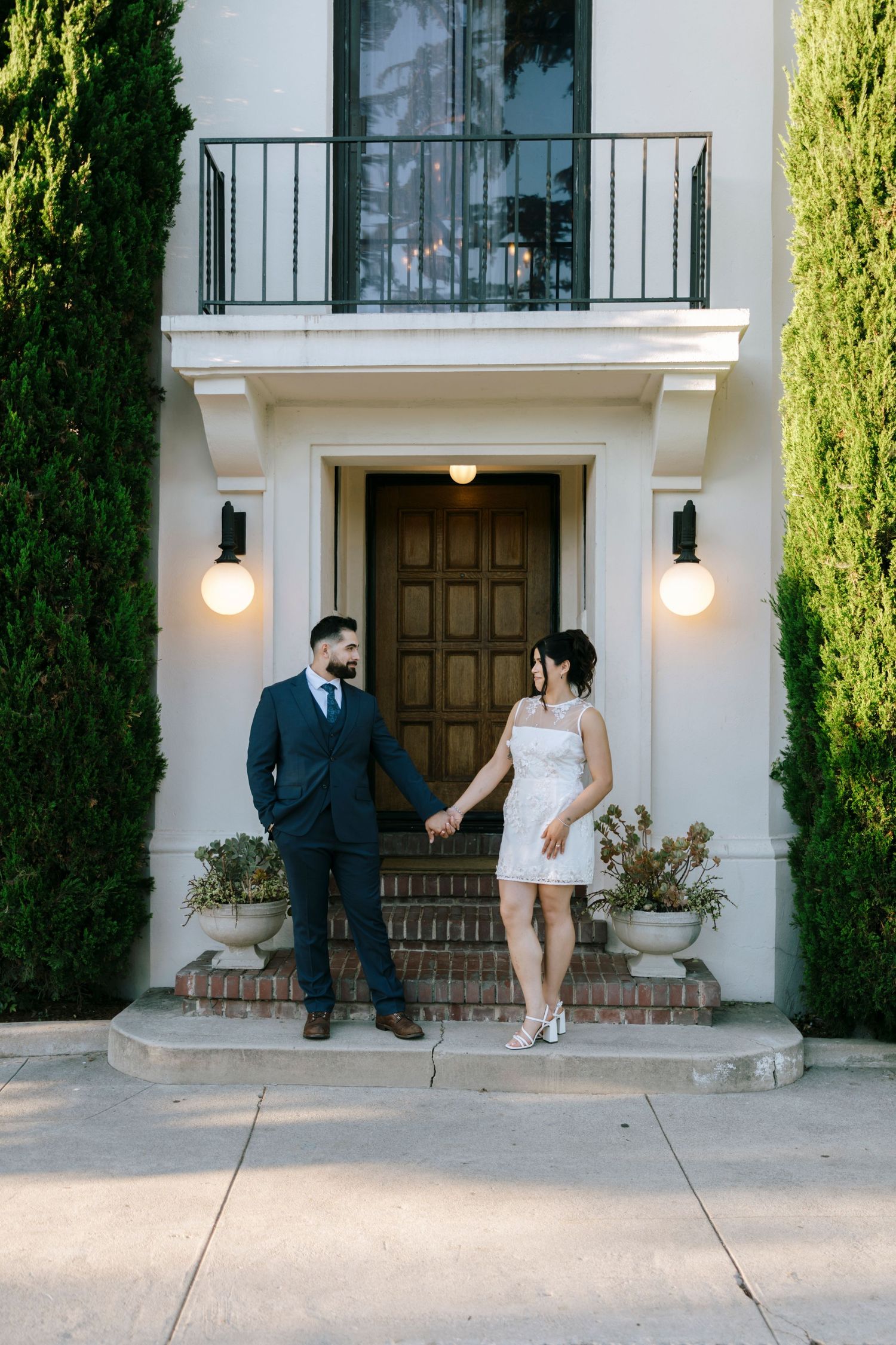 A couple poses together outside a white Mediterranean-style building with cypress trees.