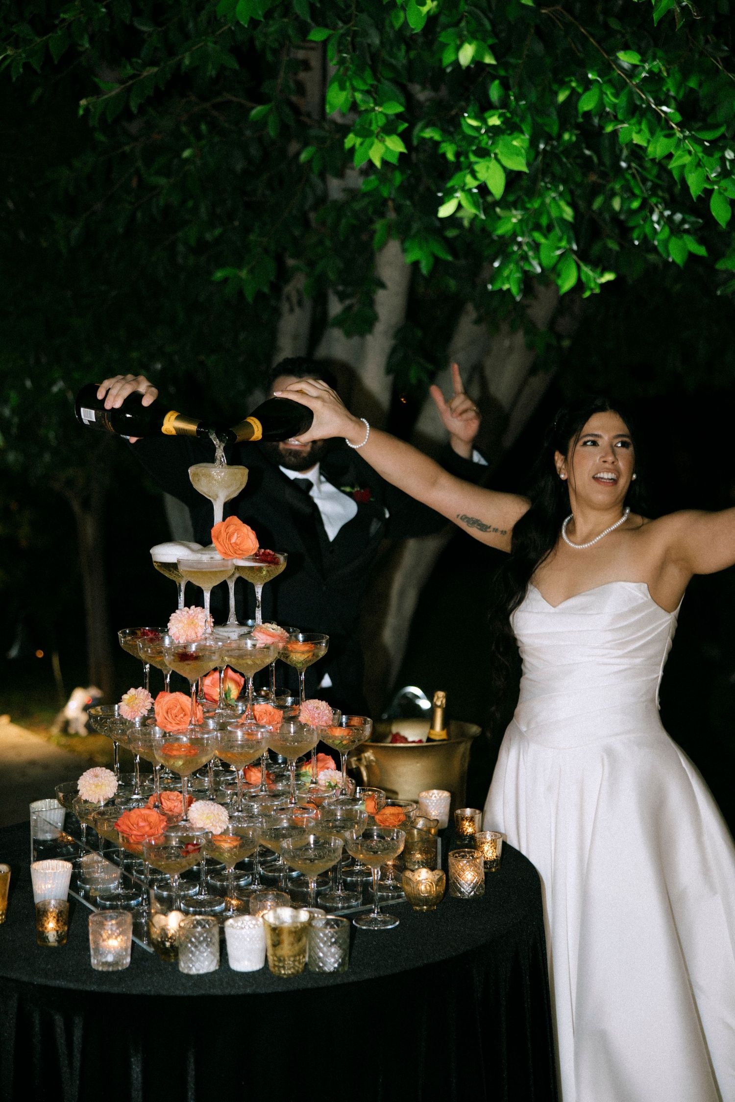 A champagne tower is being poured at an outdoor wedding reception with candlelight ambiance.