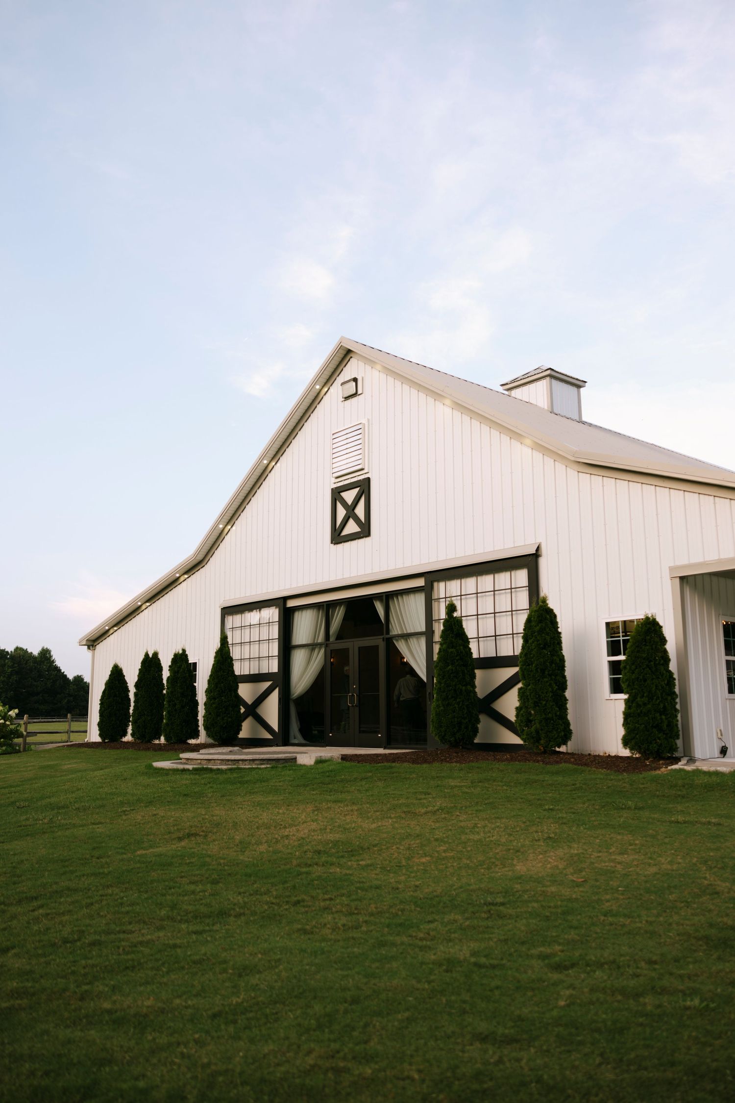 White barn wedding venue with black trim, arched entrance, evergreen trees, and lush green lawn at dusk.