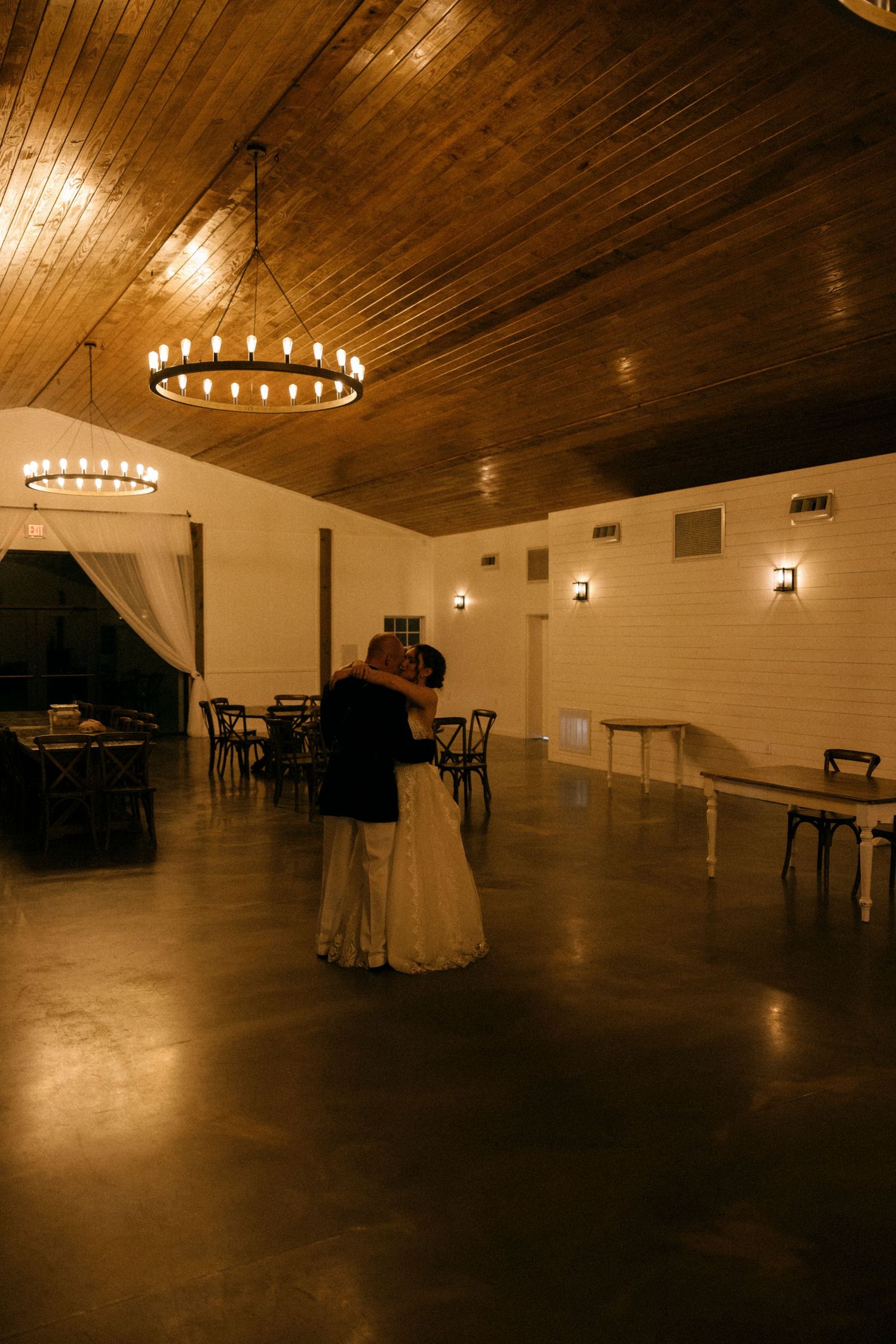 Couple sharing their first dance in a warmly lit rustic barn venue with wooden ceiling and chandelier.