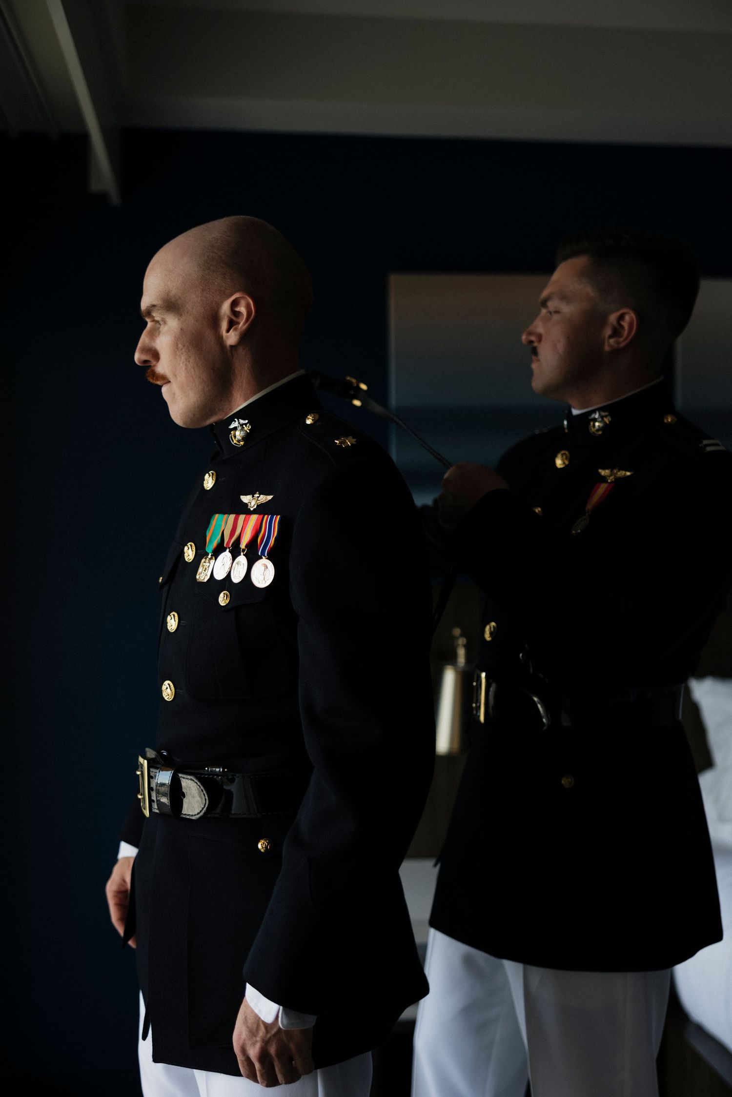 Two Marines in dress blue uniforms with medals stand at attention in a dimly lit room.