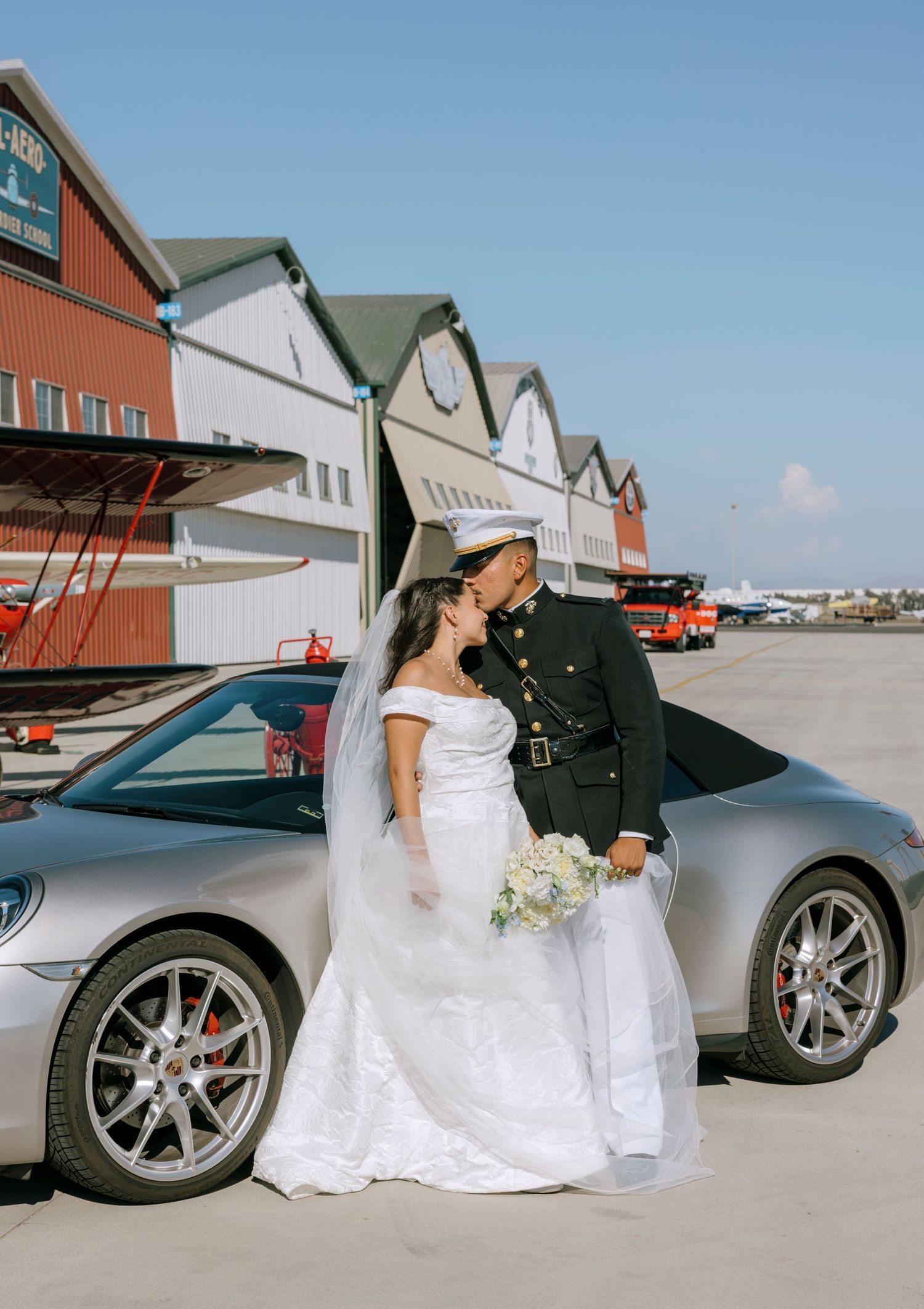 A newlywed couple poses in front of a sports car and warehouse buildings on their wedding day.