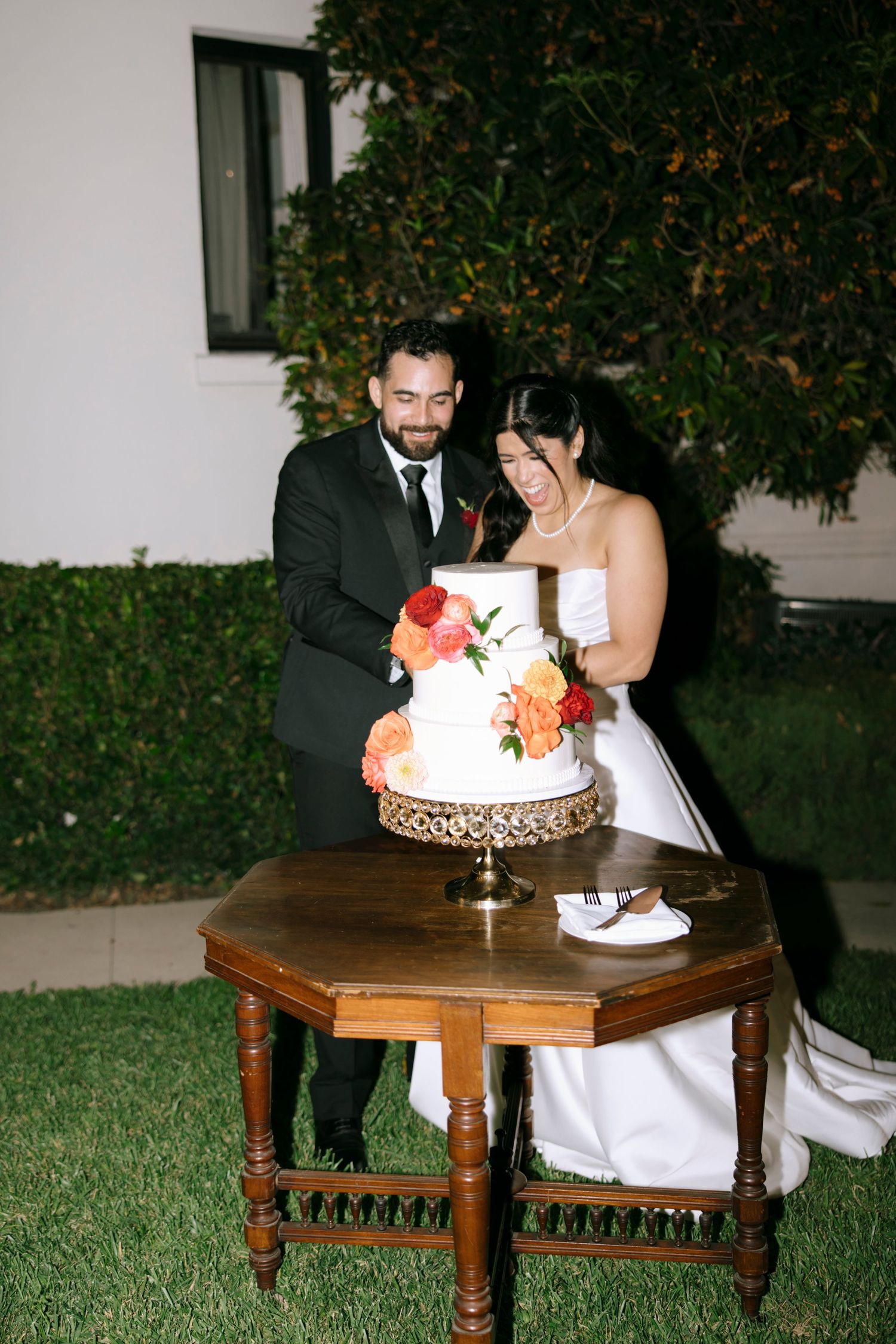 A wedding couple cuts their three-tiered coral-decorated cake on an ornate wooden table outdoors.