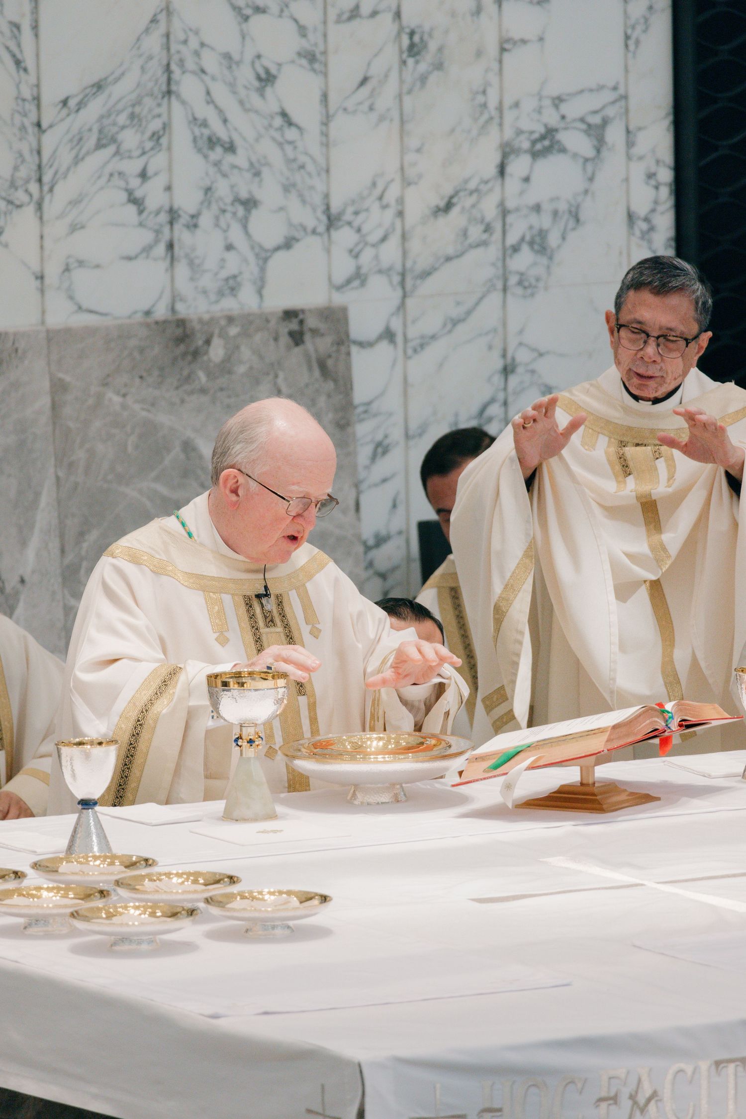 Religious leaders in cream-colored vestments seated at altar table during ceremonial service.