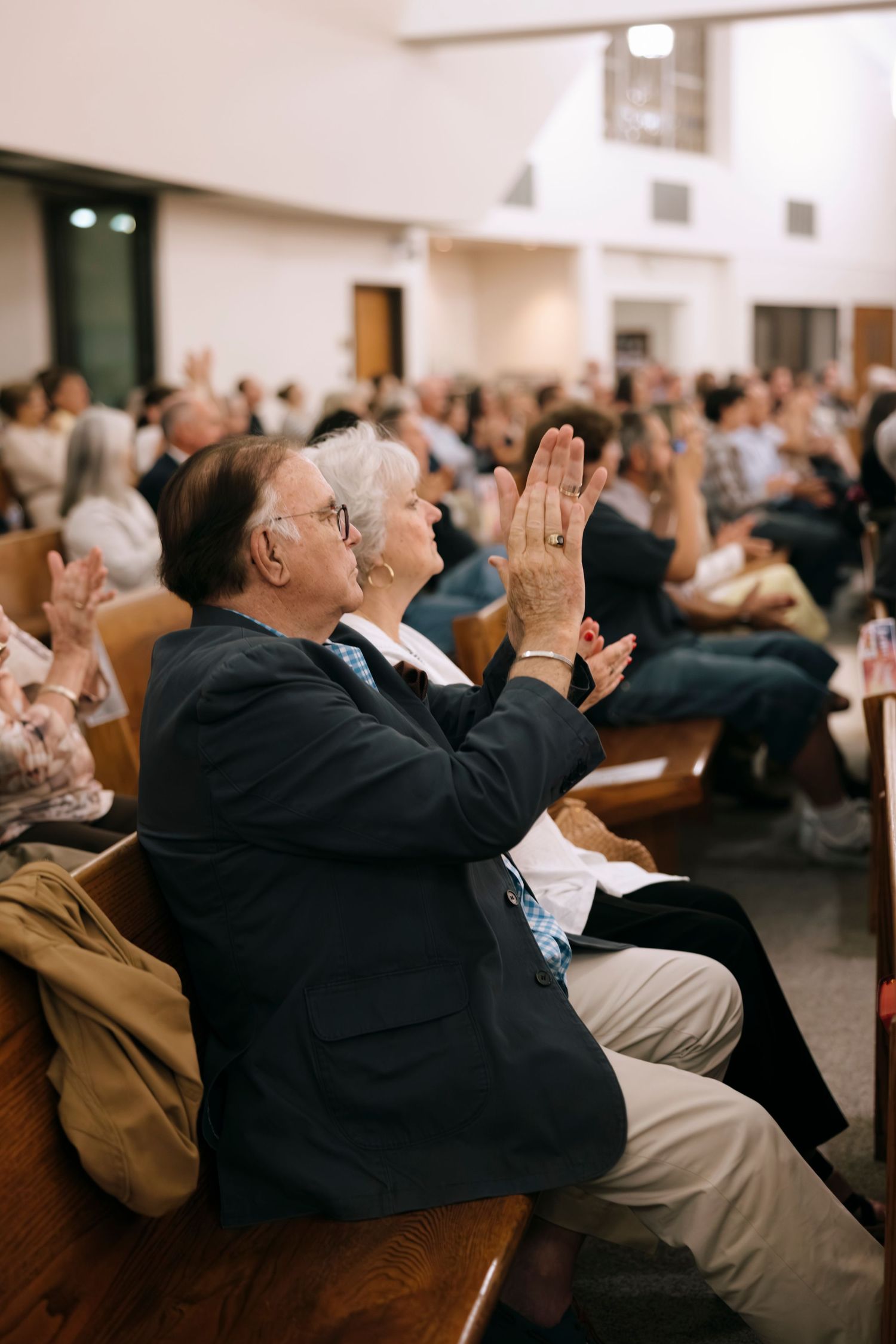 Elderly man applauds during a packed Catholic church service seated in wooden pew.