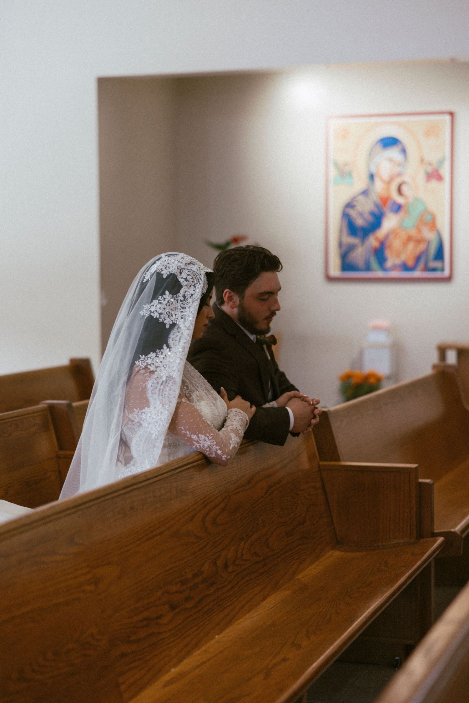 A couple sits in prayer at a church with a religious icon visible on the wall behind them.