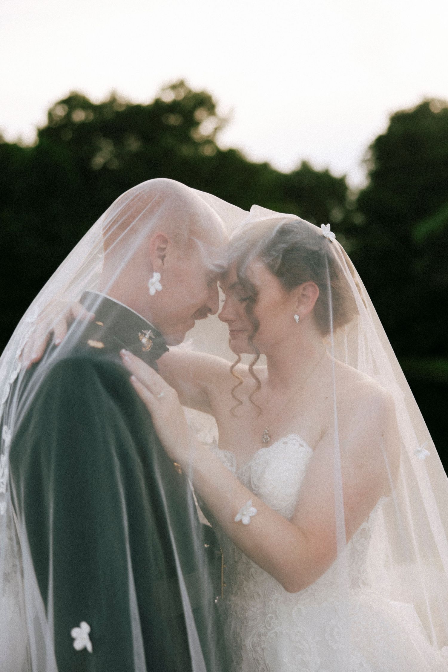 Bride and groom share an intimate moment under a sheer wedding veil, foreheads touching, surrounded by lush greenery.