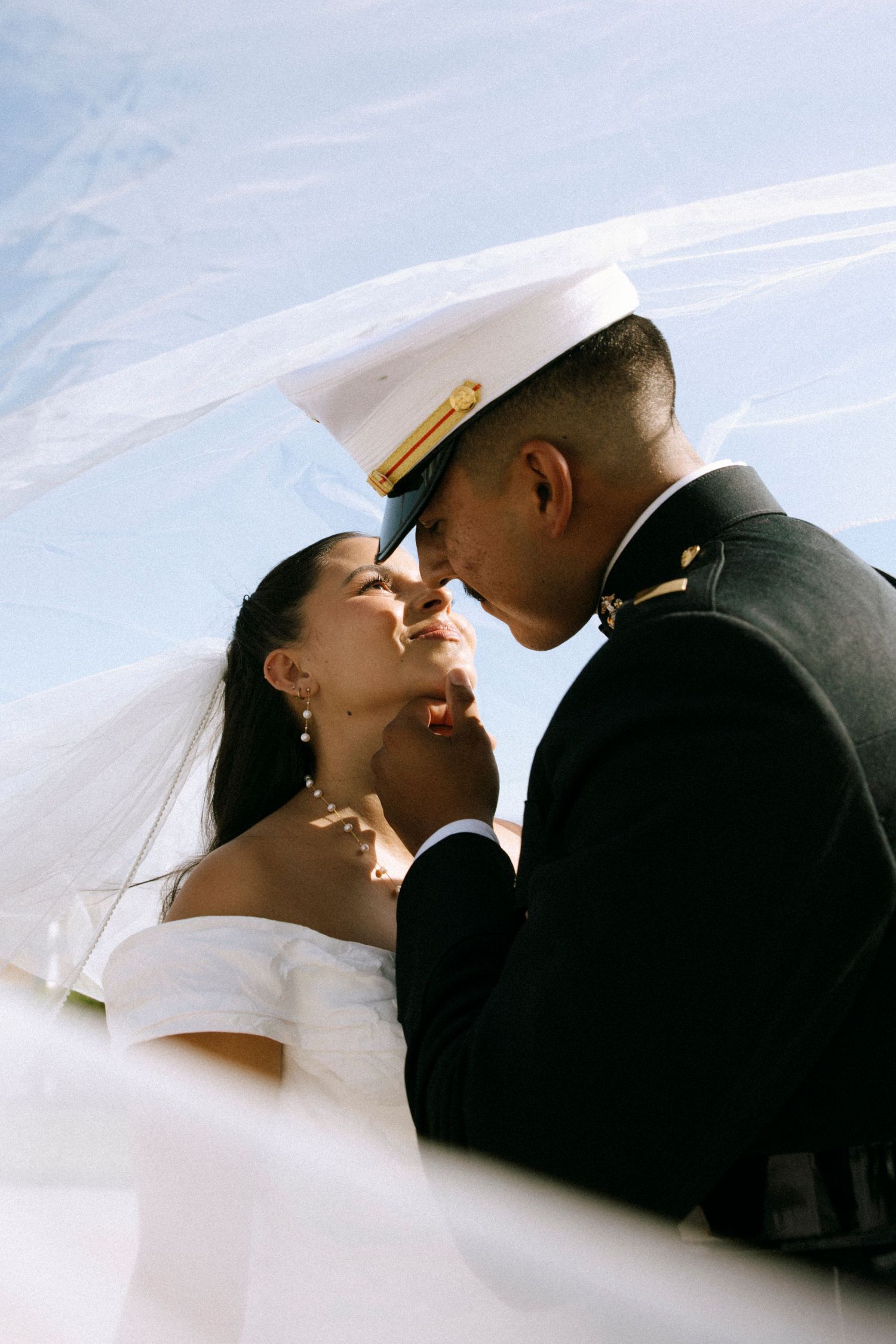 Intimate wedding portraits captured through flowing white veil against blue sky.
