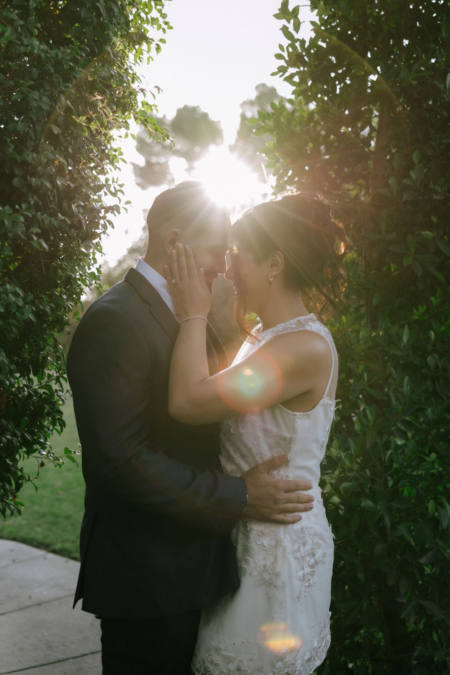 Newlyweds share tender moment in garden setting with sunlight streaming through trees.
