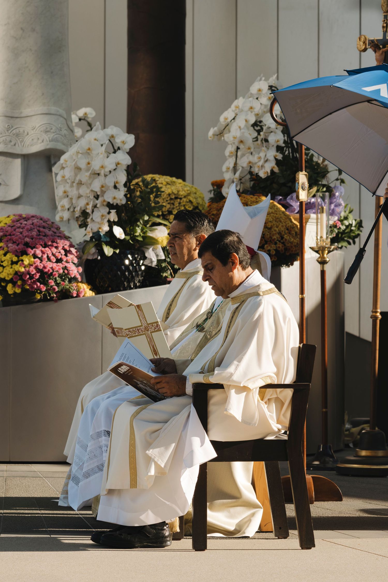 Clergy members in white robes seated on chairs surrounded by colorful floral arrangements.
