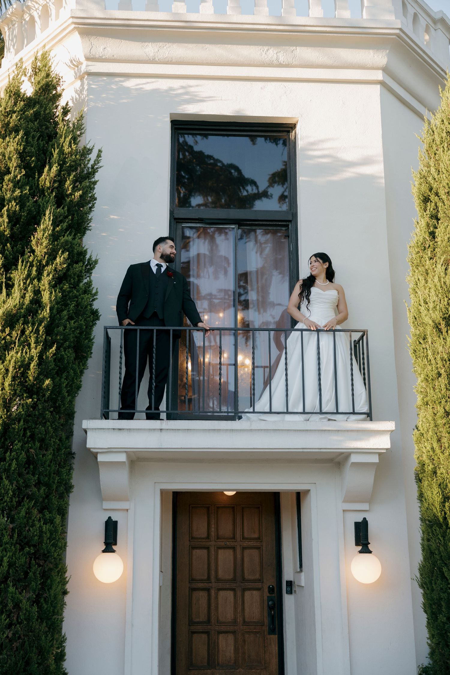 Elegant white building exterior with balcony and cypress trees framing the entrance.