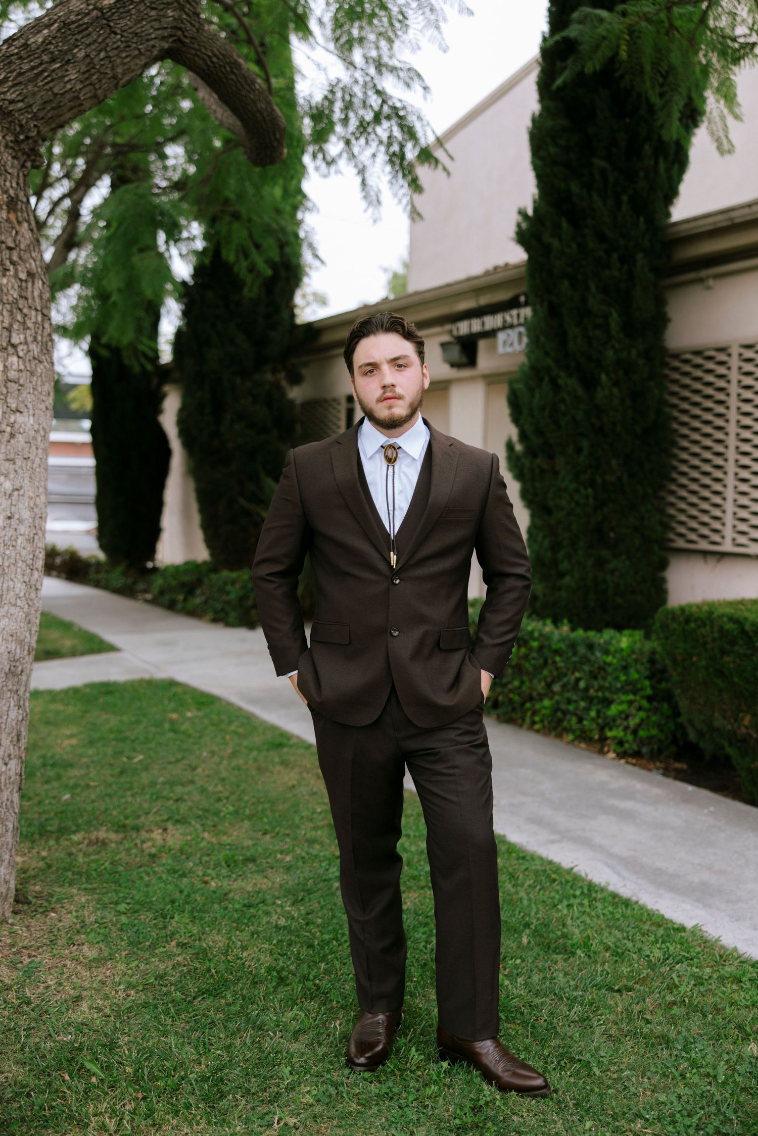 Professional in dark suit stands confidently between cypress trees on landscaped grounds.