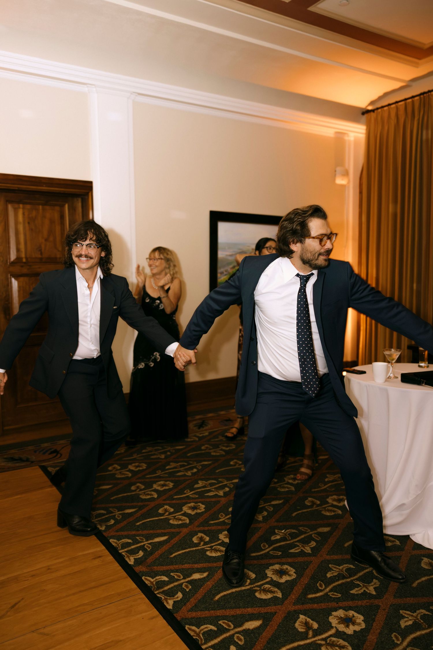 Guests perform energetic dance moves on a patterned carpet at a formal event.