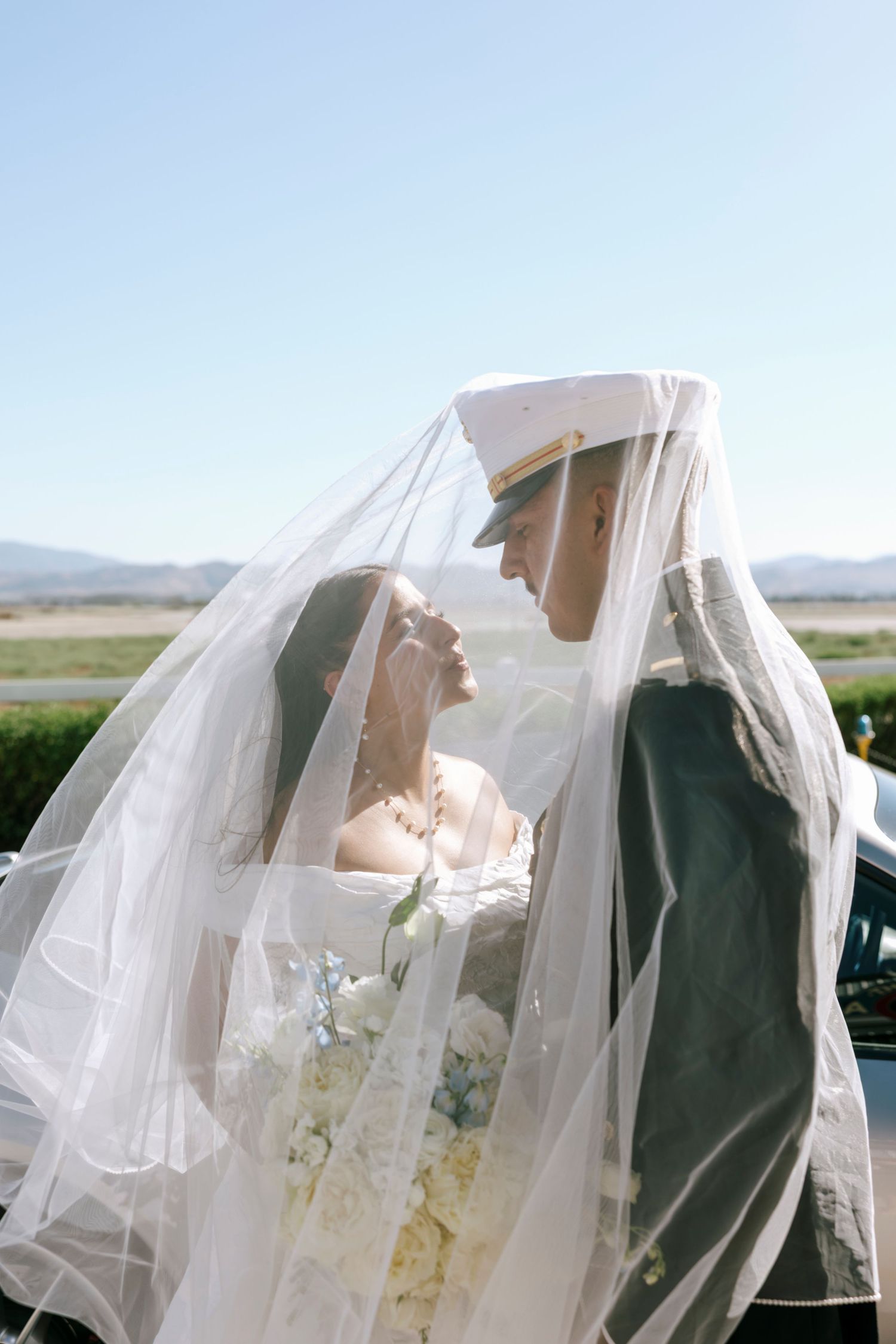 Newlyweds share an intimate moment under a flowing white veil against a bright sky.