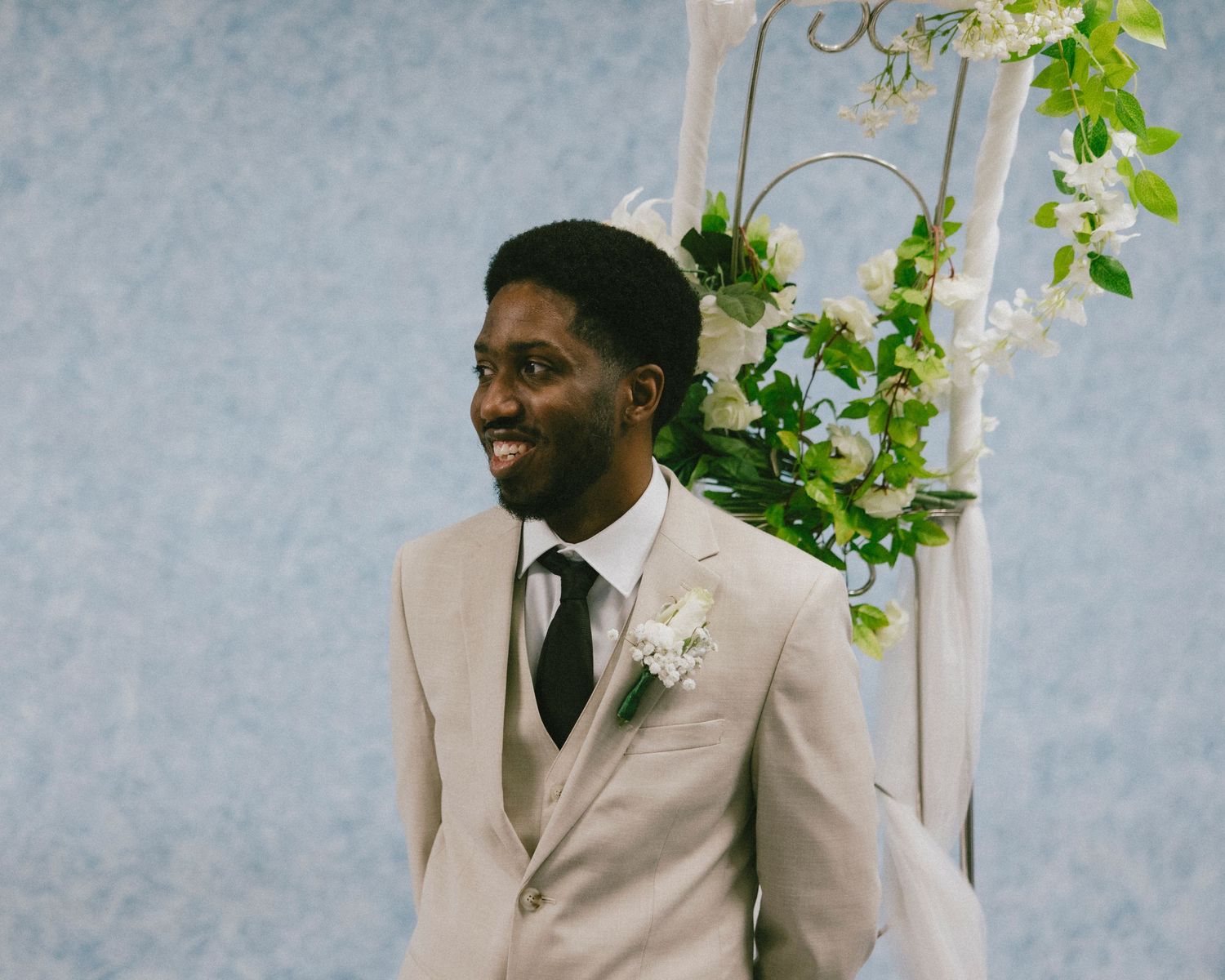 A groom in a beige suit and black tie smiles during the wedding ceremony.
