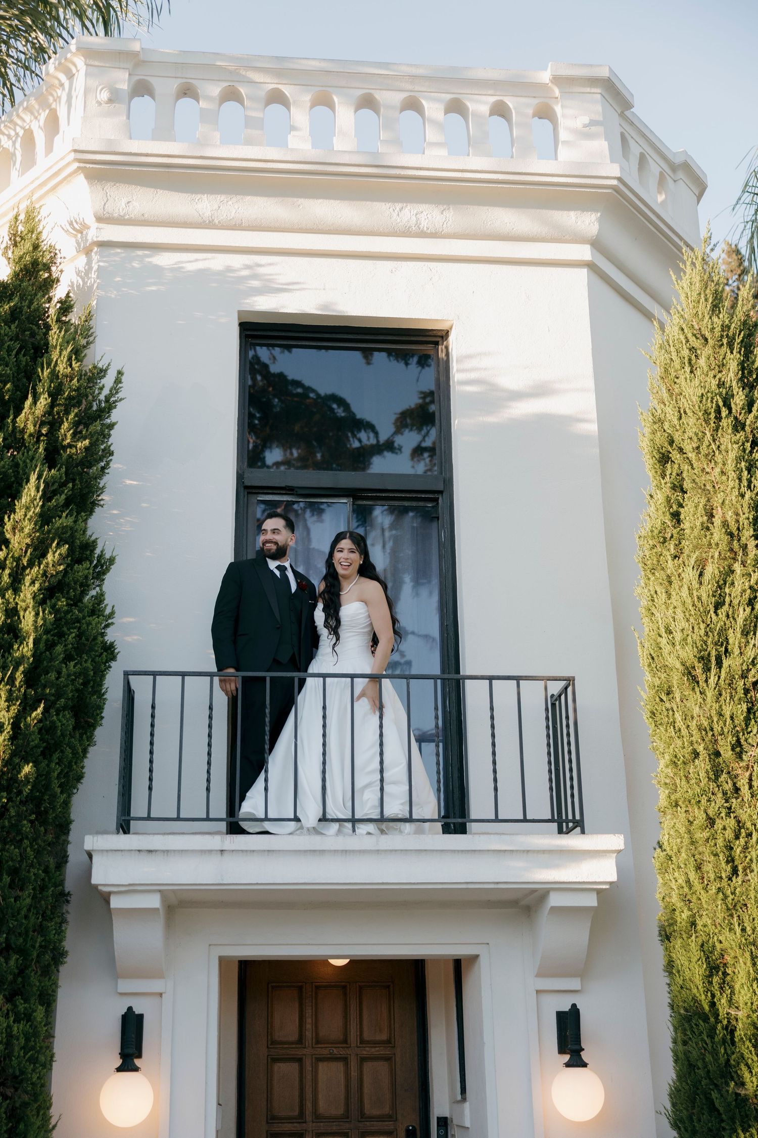Couple standing on a white Mediterranean-style balcony with ornate architectural details.