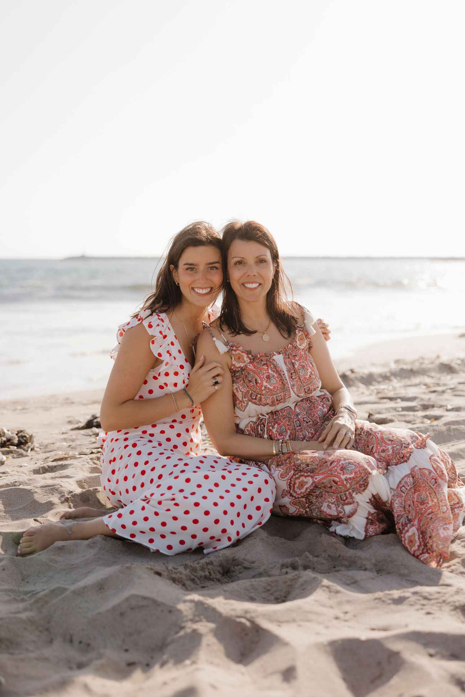 Two women in red and white outfits sitting together on a sandy beach, smiling at the camera.