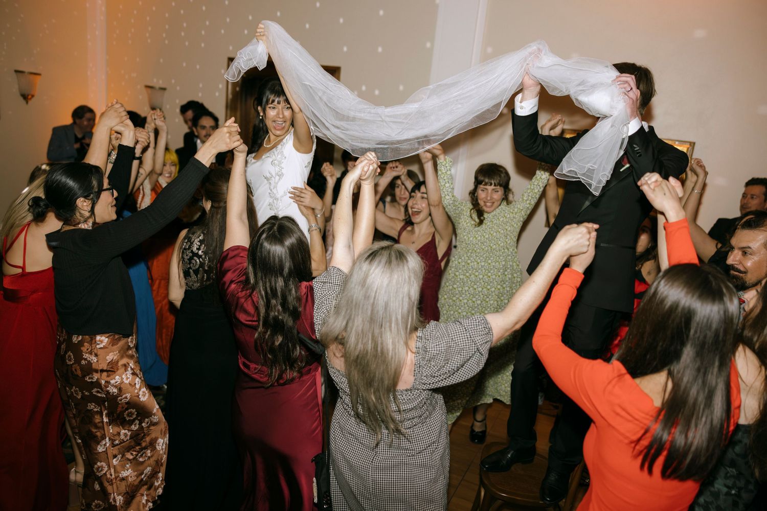 Celebration scene with guests raising white fabric while dancing at an indoor event.
