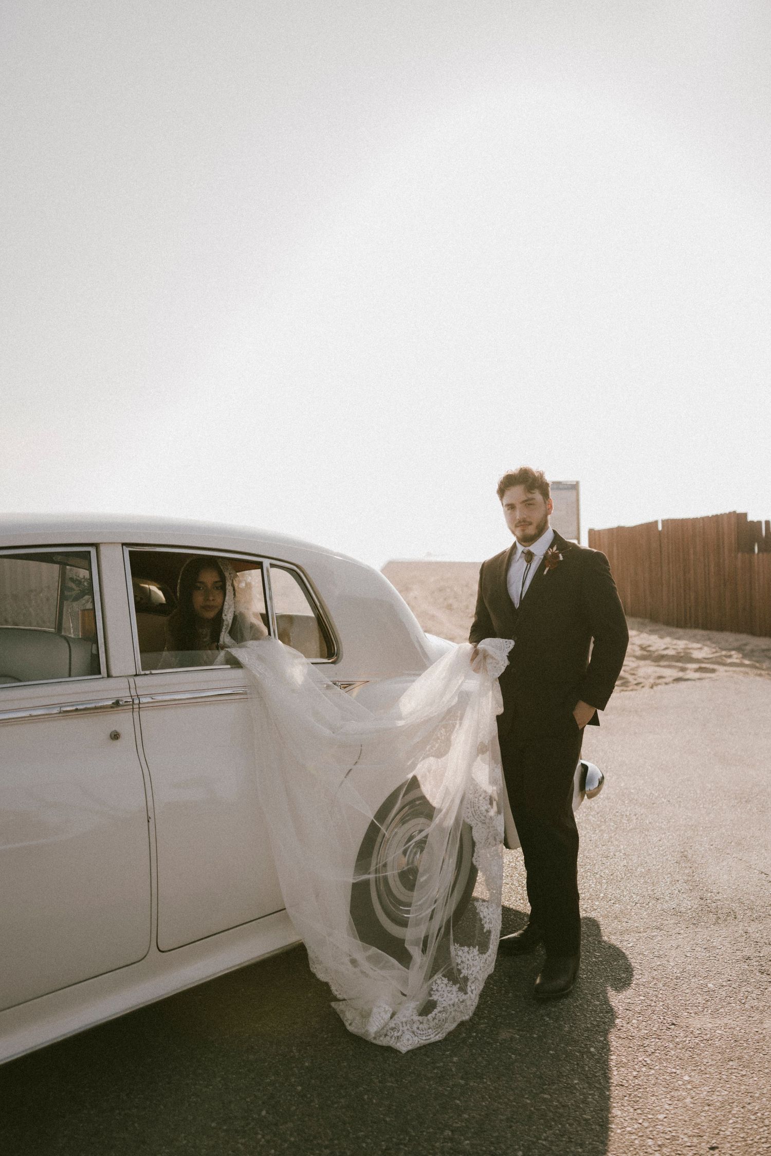 Dramatic wedding photos with flowing veil and classic white car in desert setting.