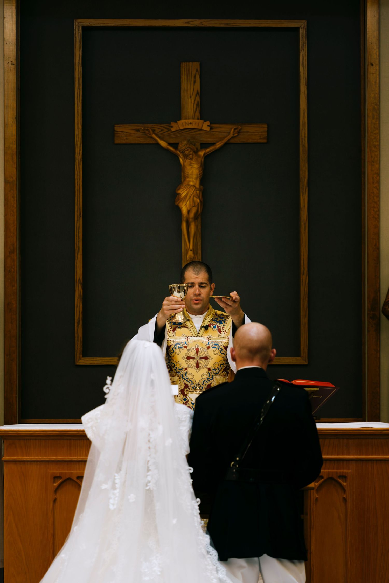 Priest elevating chalice during Catholic wedding ceremony at church altar with crucifix, bride in white gown and groom in black suit.