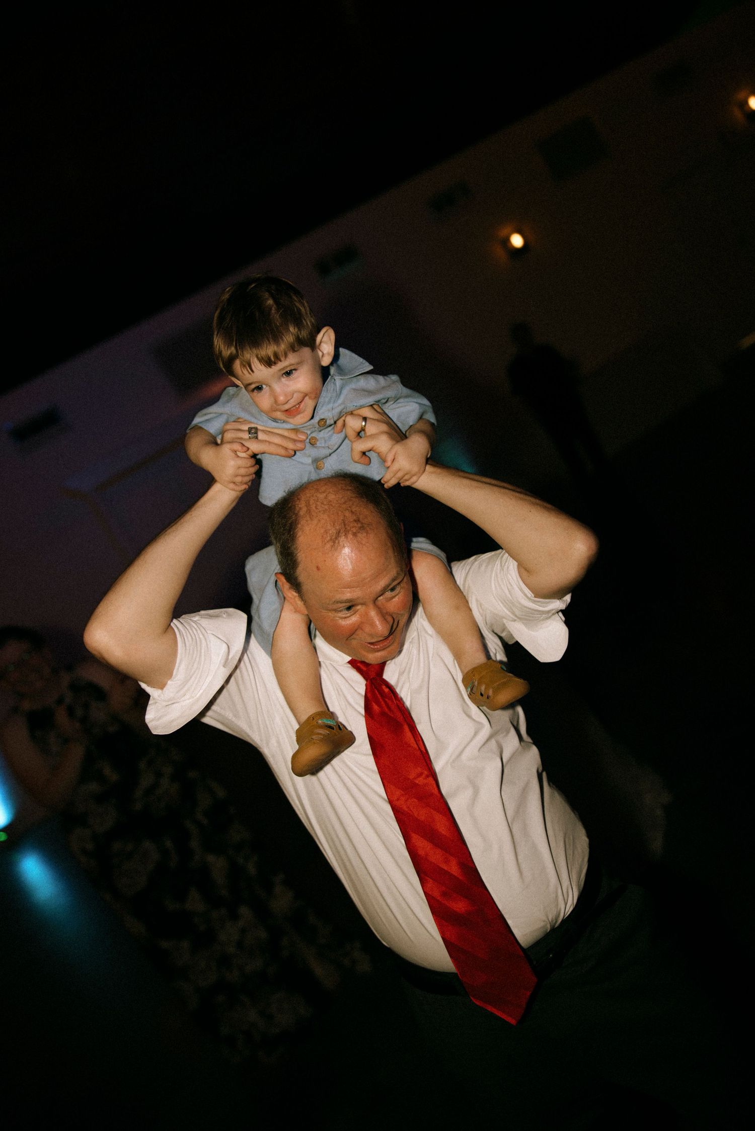 A bald man in a white shirt and red tie carries a young boy on his shoulders at a dark event venue.