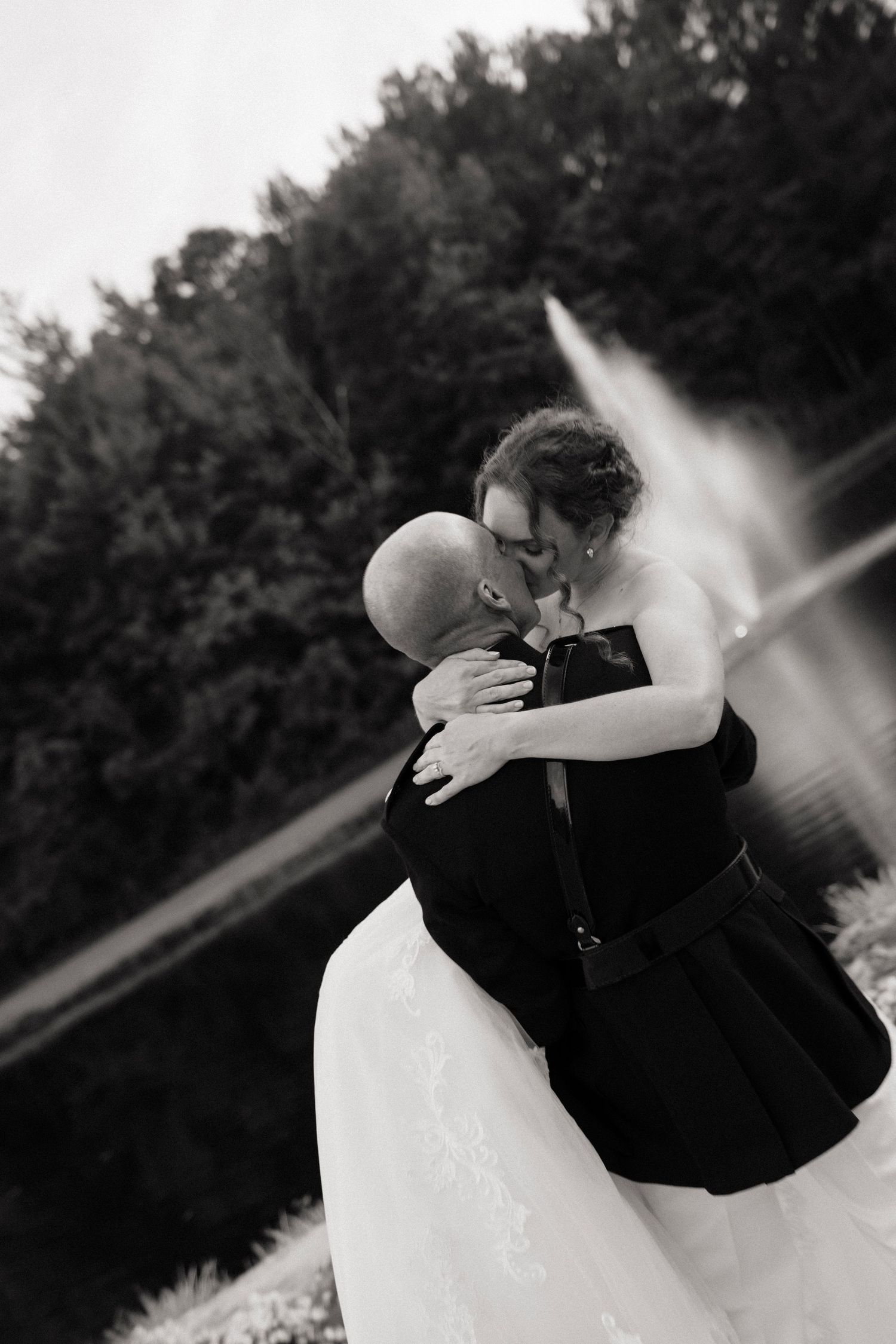 Black and white photo of a bride and groom sharing a kiss near a fountain surrounded by lush trees.