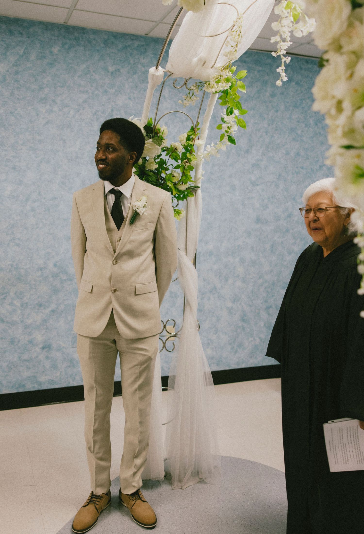 A groom in a light beige suit stands under a floral arch during a wedding ceremony.
