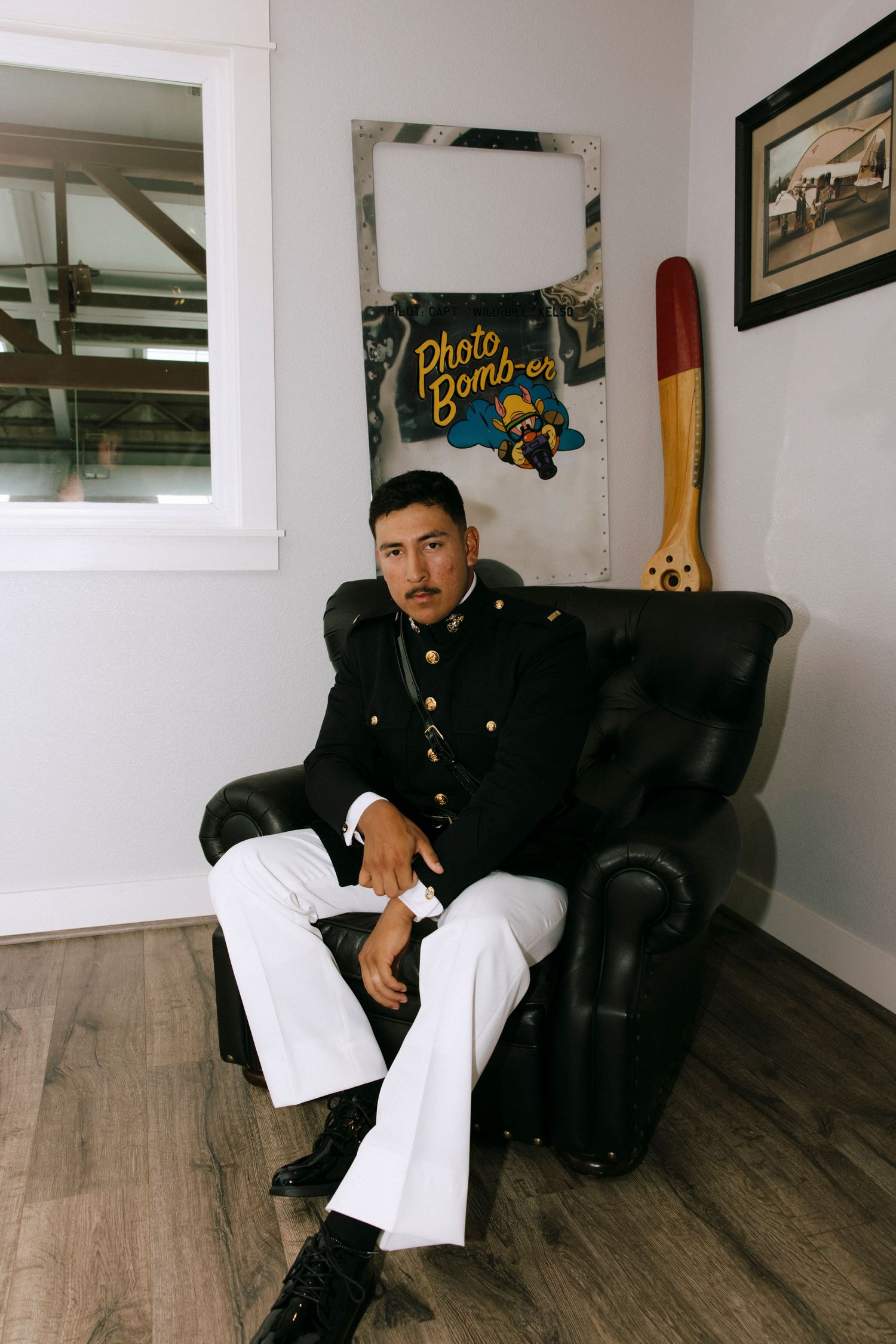 A person in military dress uniform sits casually on a dark leather couch in a room with wood flooring.