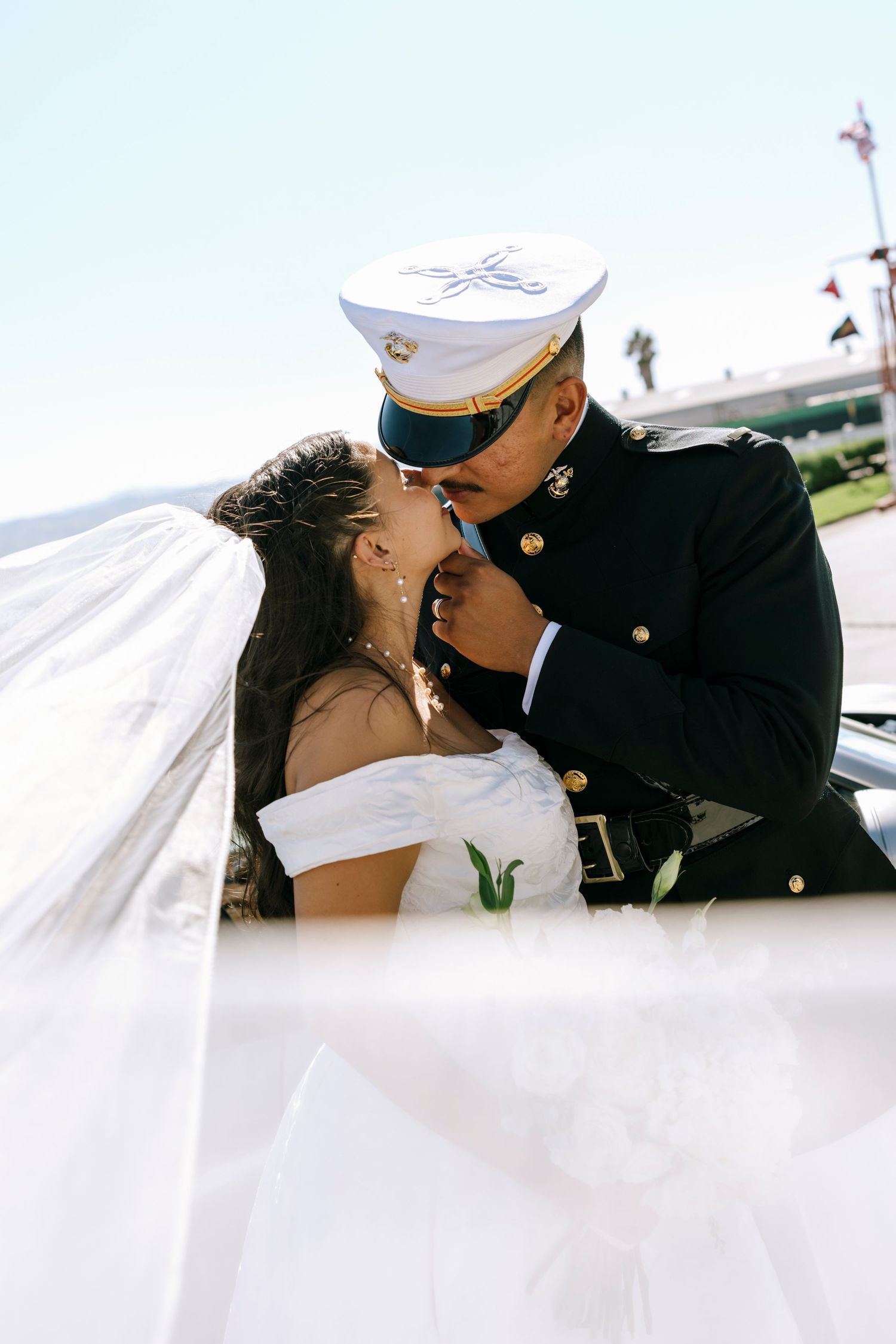 A romantic wedding kiss between a couple with flowing veil and military dress uniform.