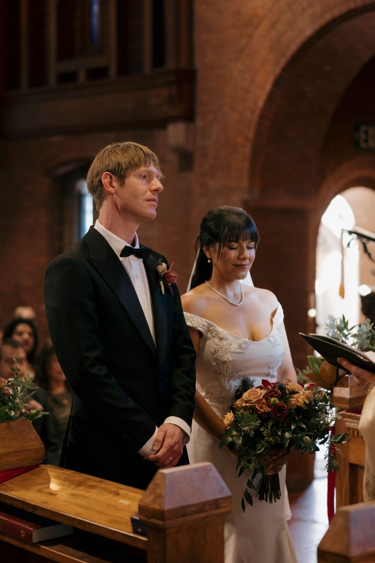 Couple standing at wooden church altar during wedding ceremony with floral bouquet.