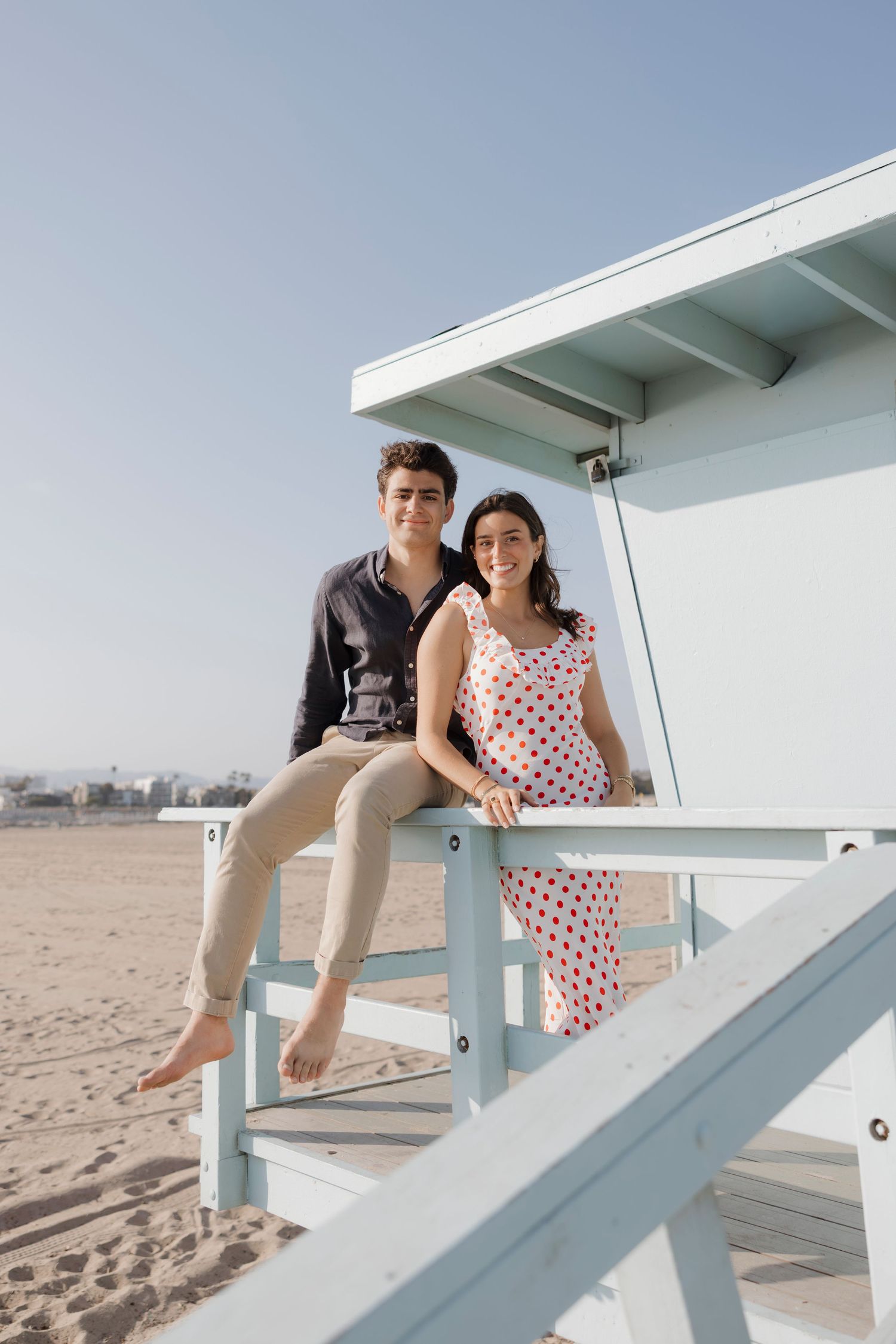 Young couple smiling on a lifeguard stand at a sandy beach on a clear day.
