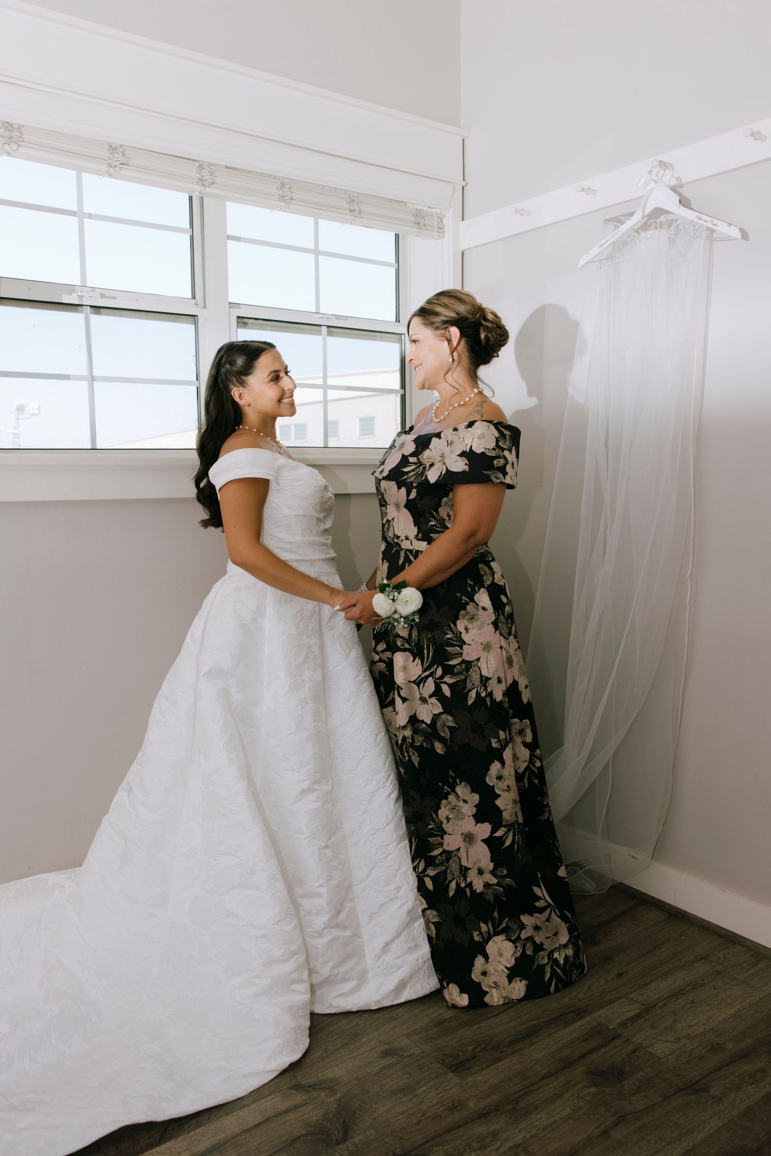 An emotional moment between a bride in white and a guest in a floral dress near a window.