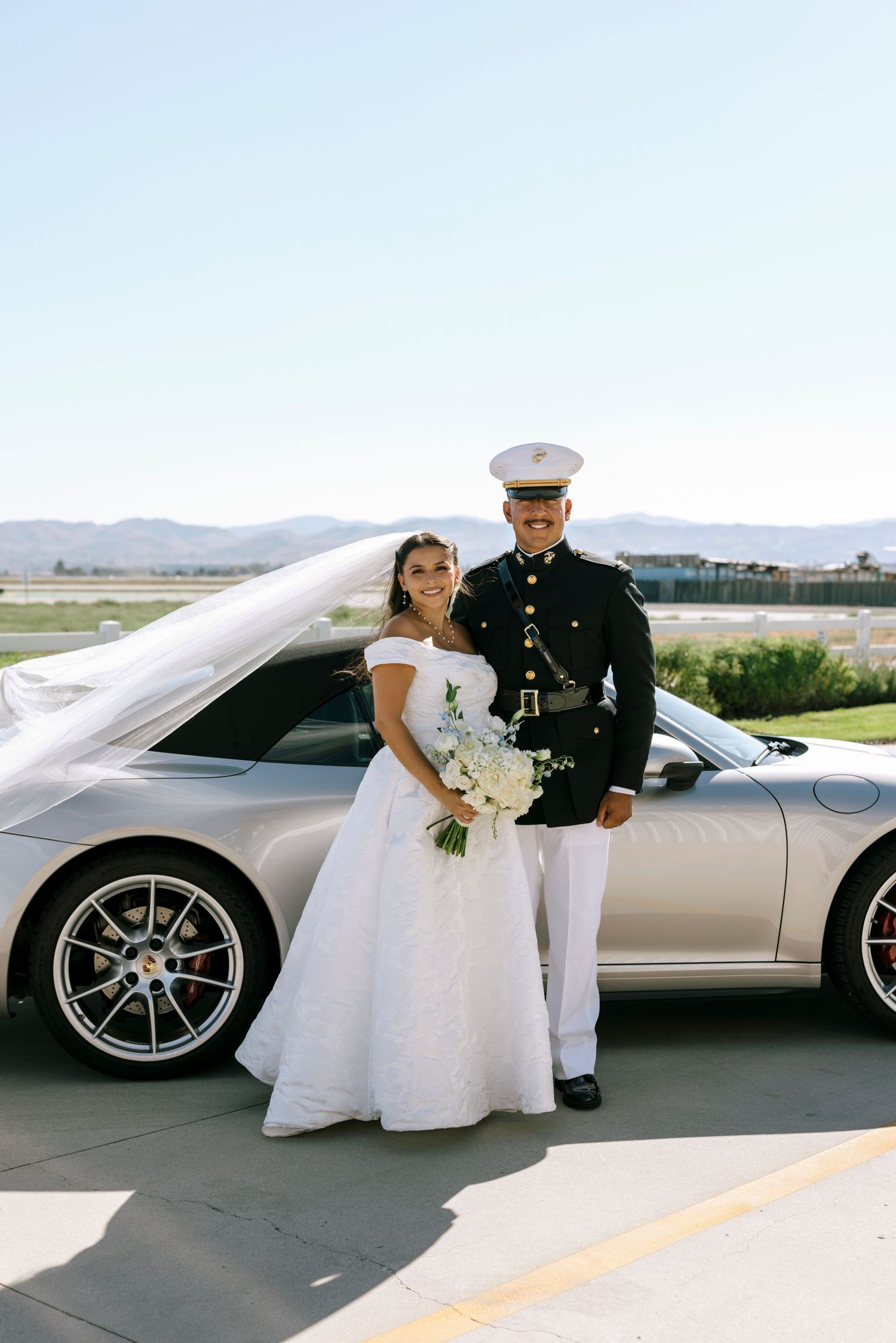 A military wedding couple poses with a silver sports car on a sunny day.