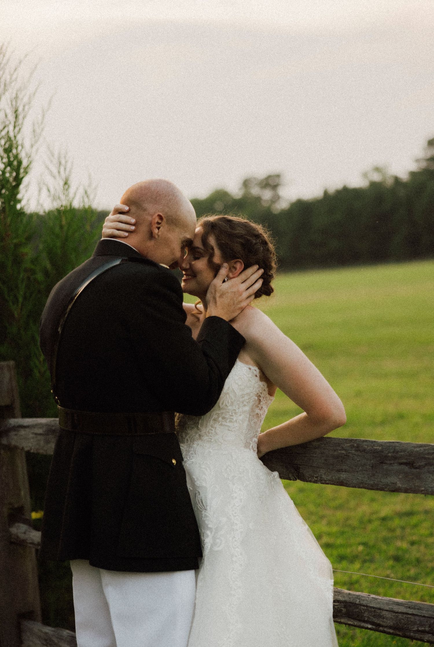 A bride and groom share a romantic forehead touch by a wooden fence in a lush green outdoor wedding setting.